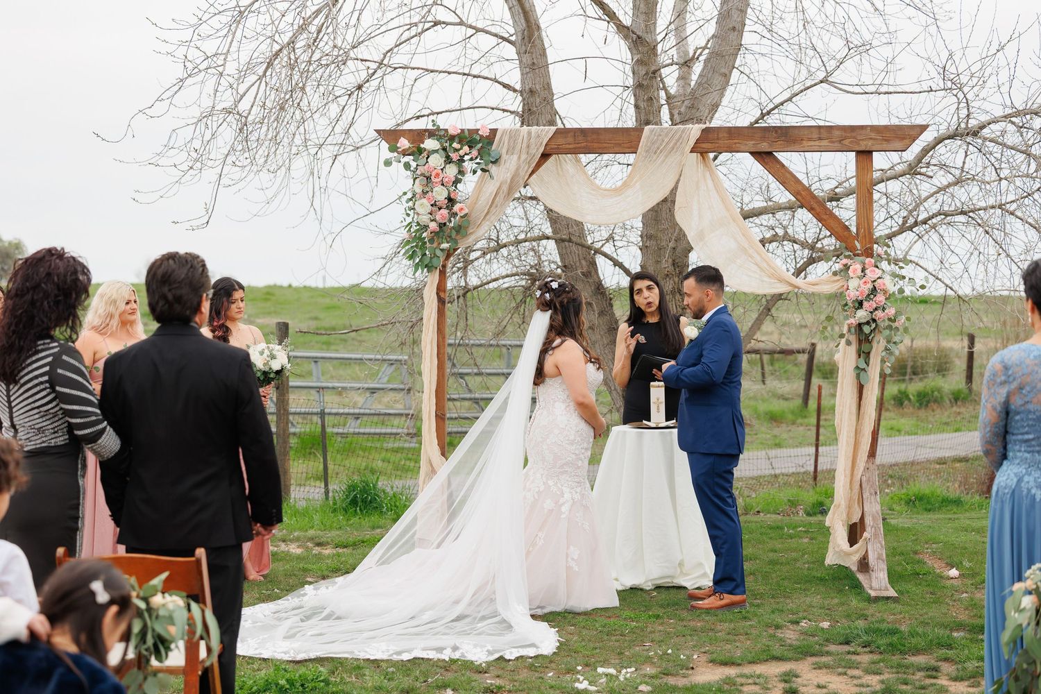A wedding ceremony takes place under a rustic wooden arch decorated with floral arrangements in an outdoor countryside setting.