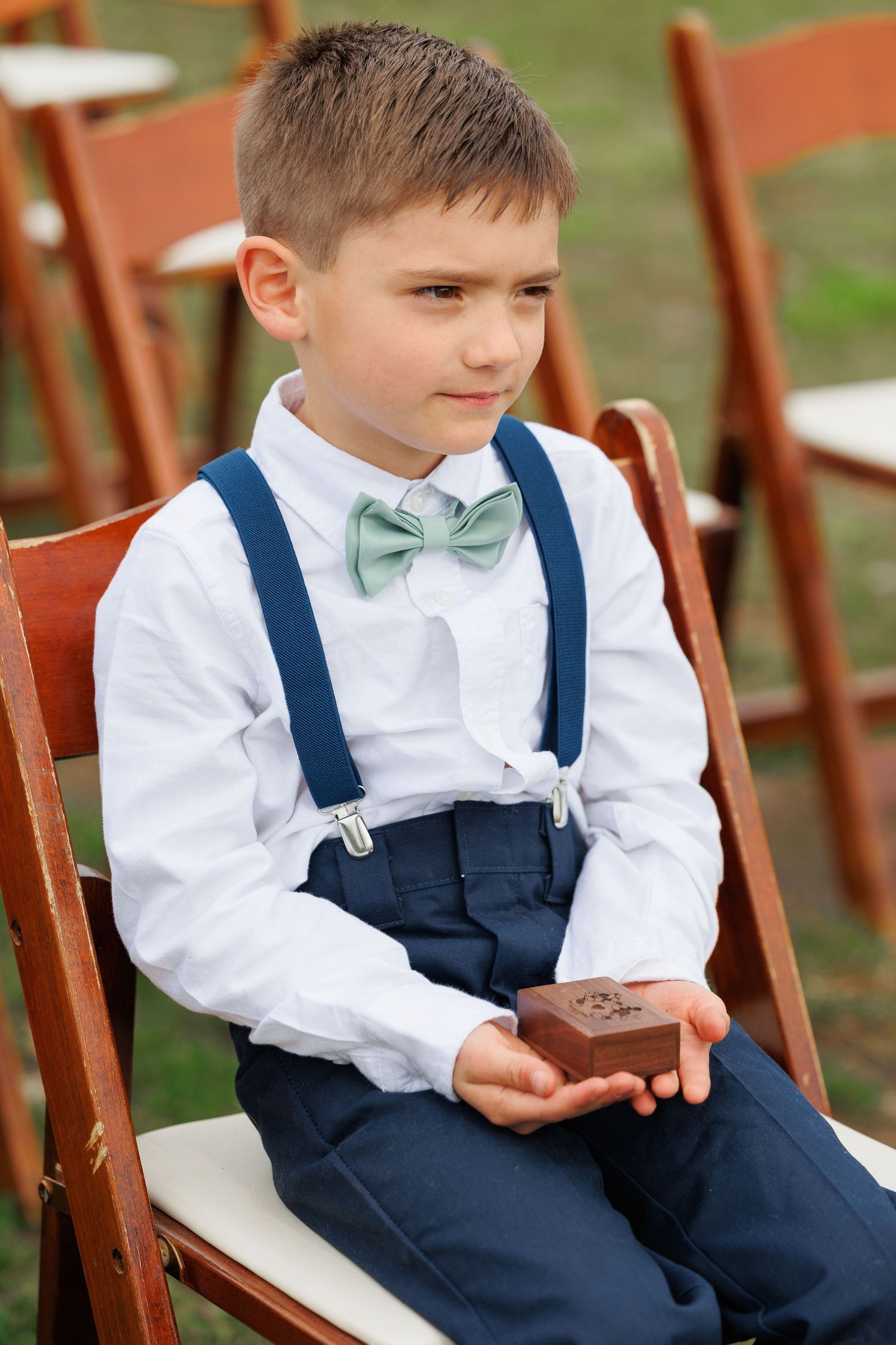 Young ring bearer in navy suspenders and mint bow tie sits on wooden chair at outdoor wedding ceremony.
