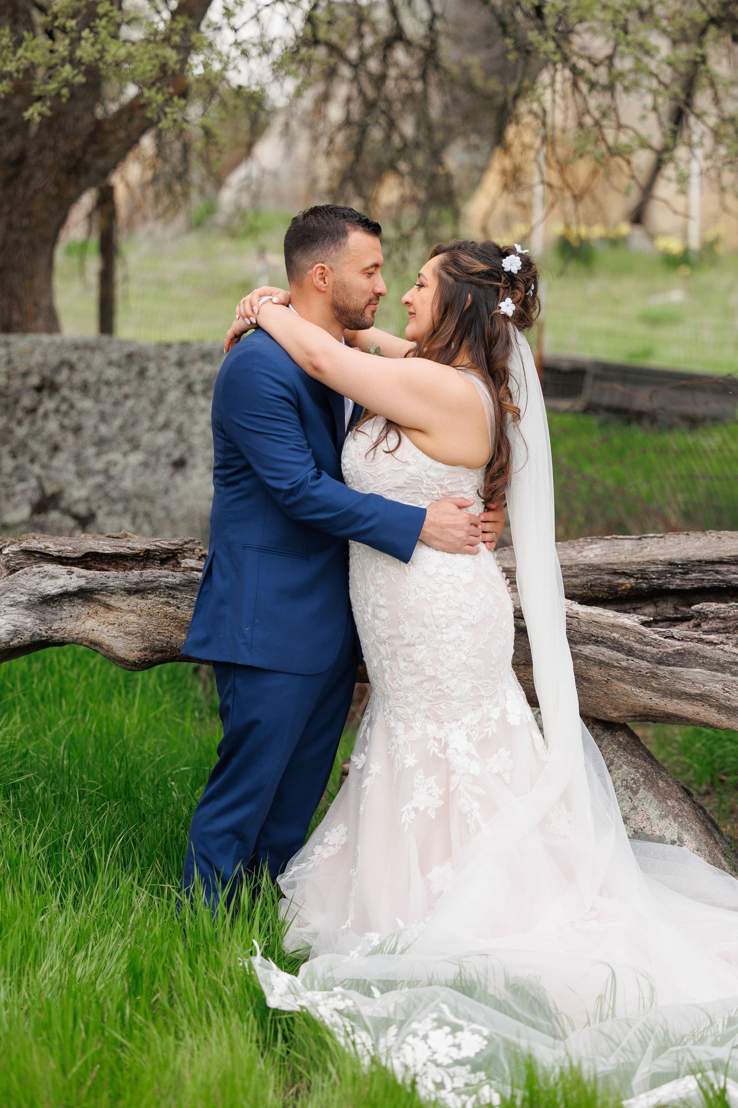 A romantic wedding couple embraces near a fallen tree trunk in a lush green meadow during their outdoor photo session.