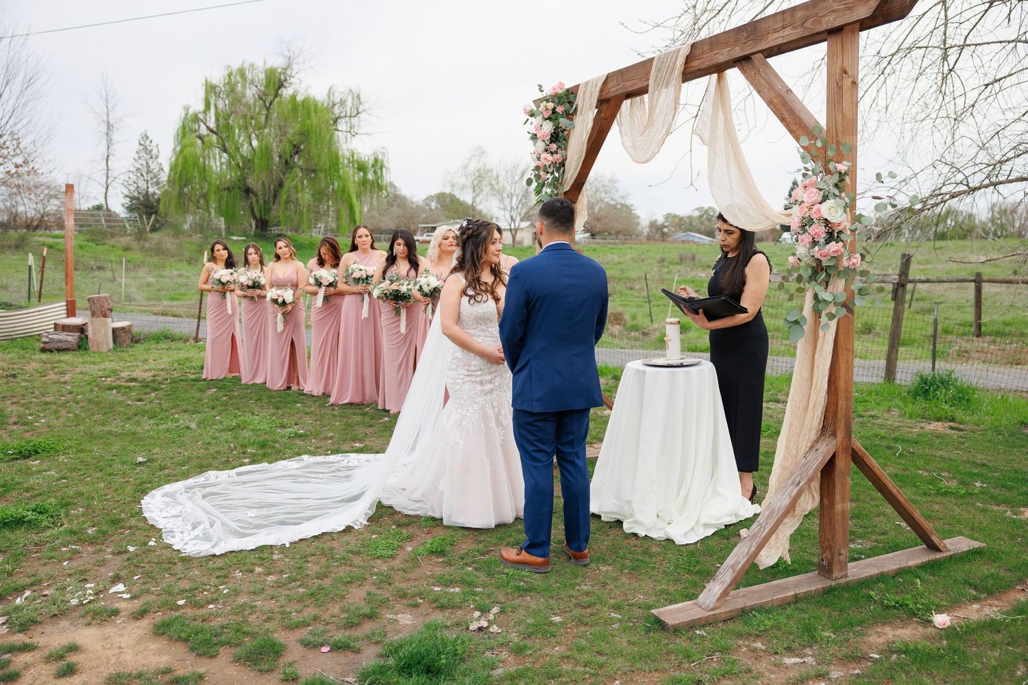 Wedding party gathers under a rustic wooden arch in an outdoor garden setting during spring.