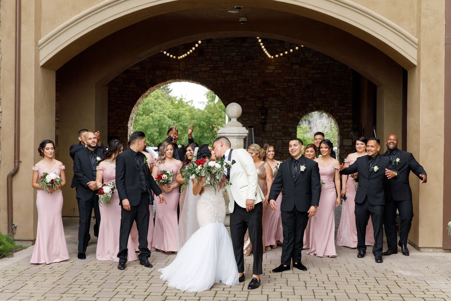 A wedding party in formal attire stands in a row under an elegant stone archway with pink and white decor.