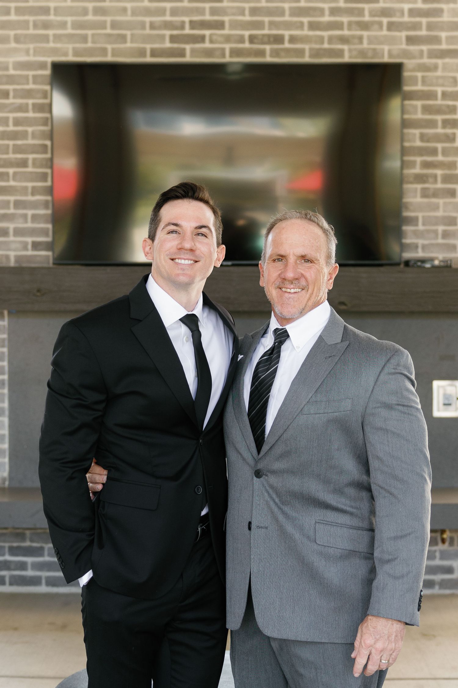 Two people in formal attire pose together in front of a brick wall, one wearing a black suit and one in gray.