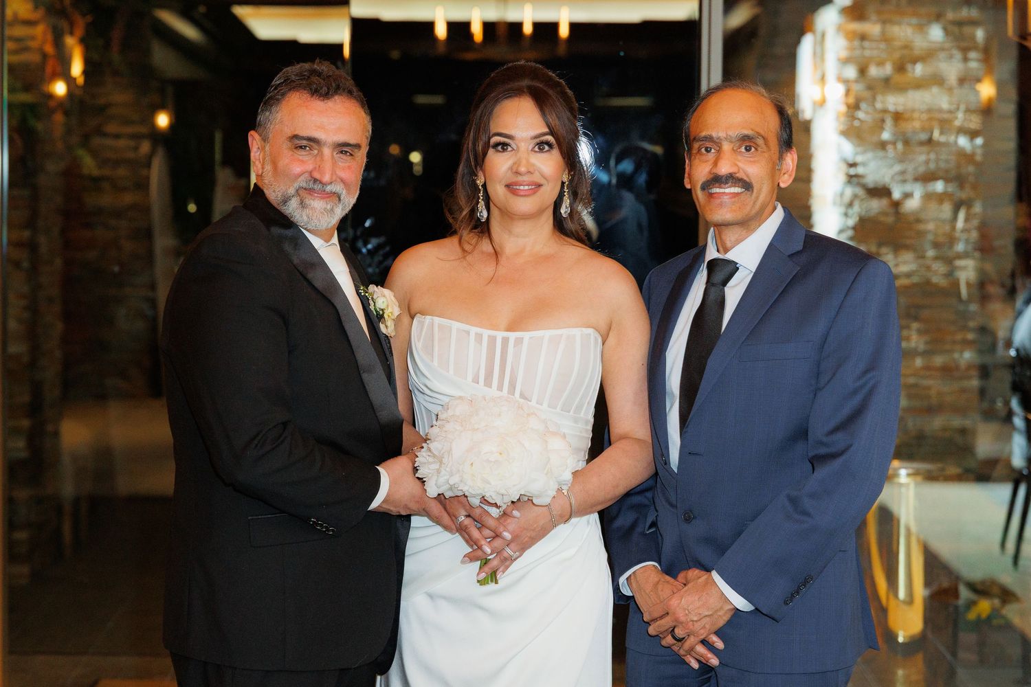 Wedding party members pose together in formal attire at an indoor venue with brick walls.