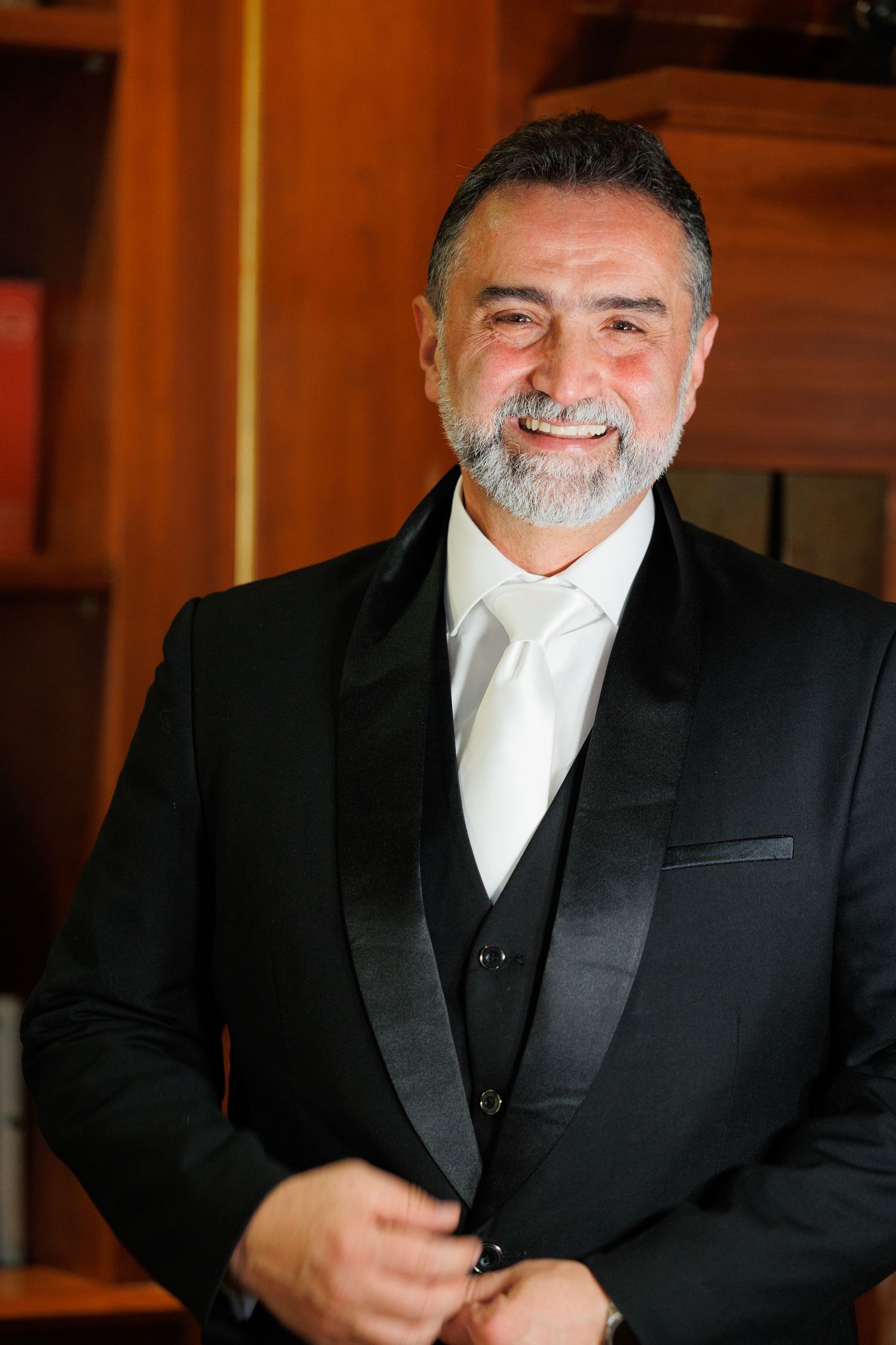 A bearded man in a formal black tuxedo with white shirt and vest smiles warmly in an elegant indoor setting.