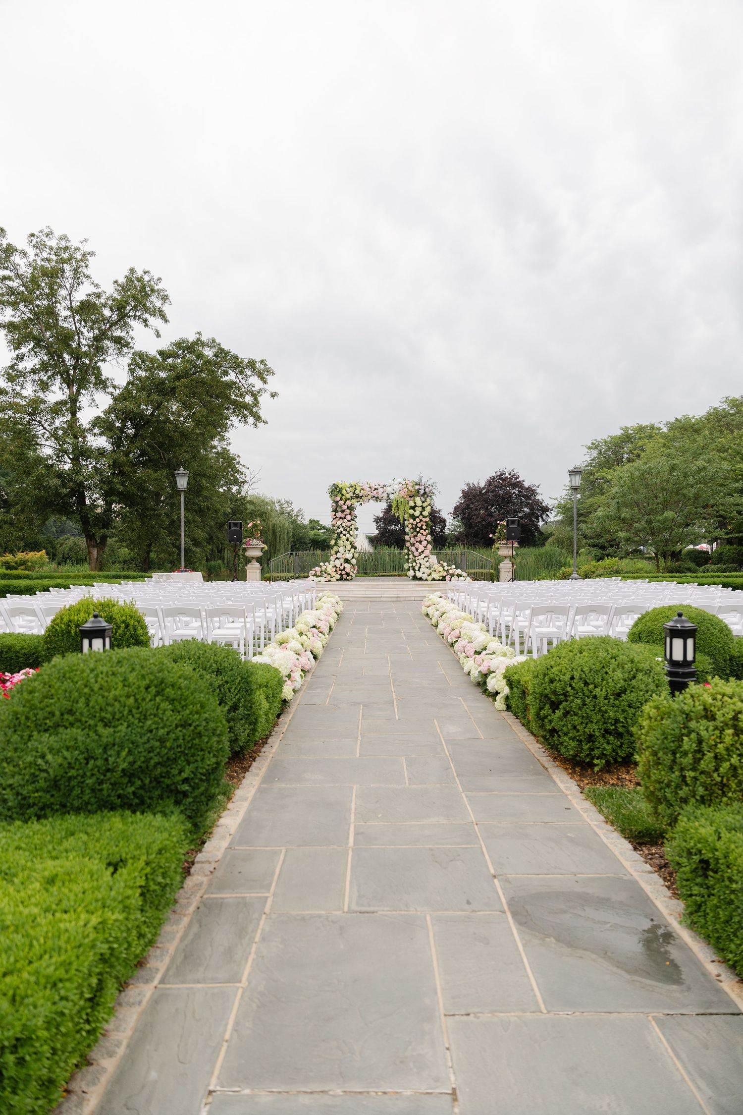 An outdoor wedding ceremony setup with white chairs lined along a stone pathway leading to a floral arch.