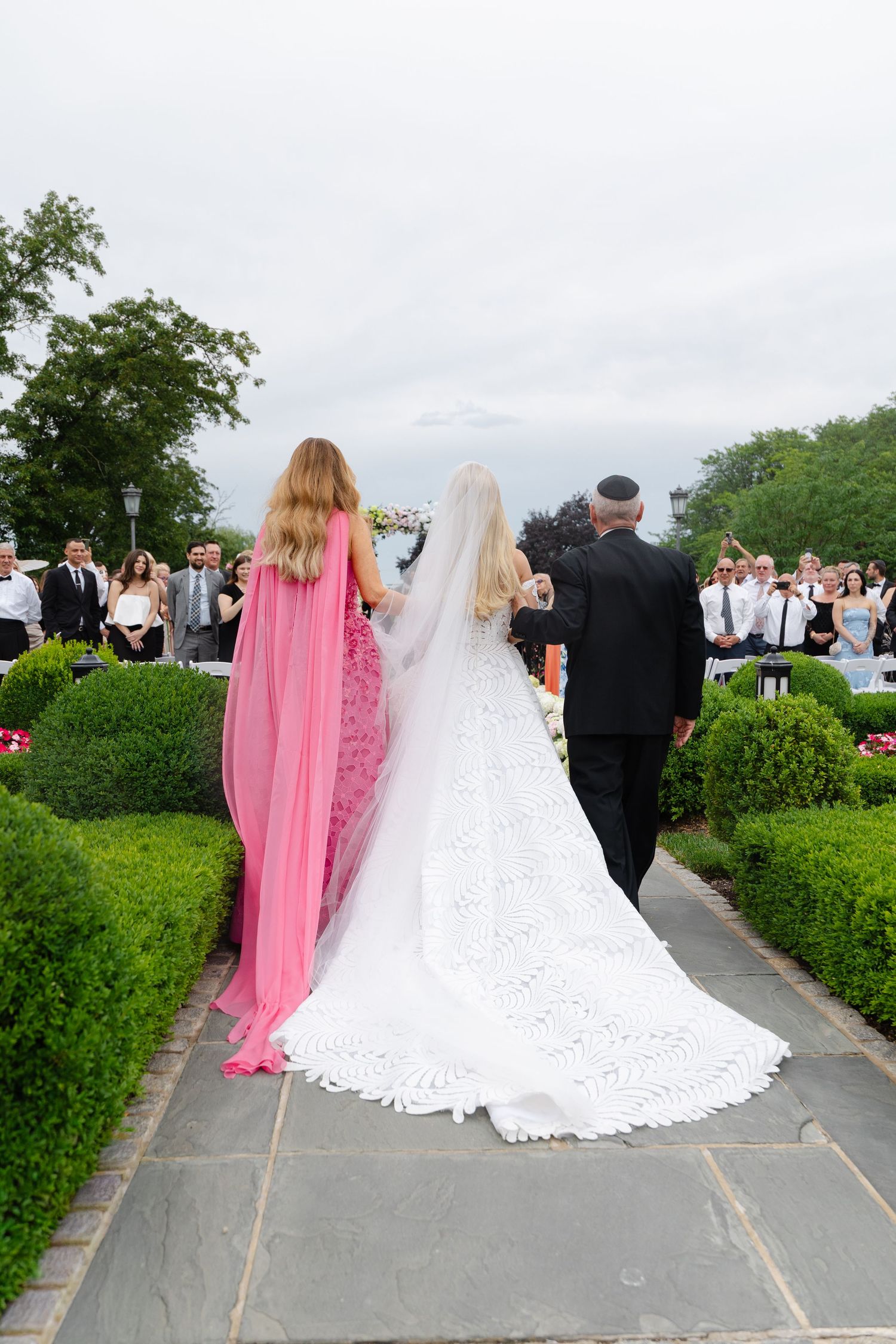 Wedding party walking down stone path with bride in white gown and bridesmaid in pink dress at outdoor garden ceremony.