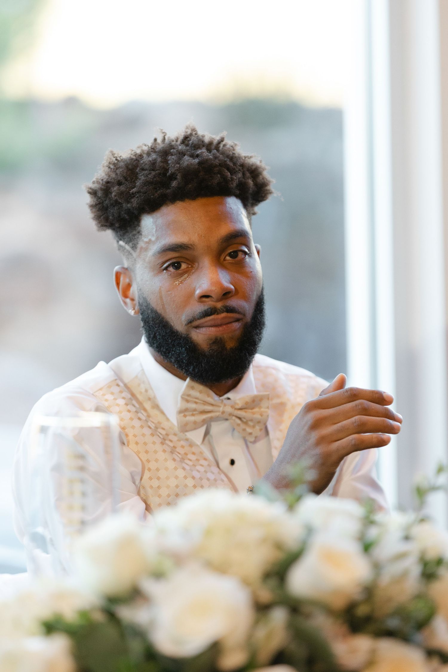 A groom in a white polka dot bow tie and jacket sits thoughtfully at a wedding reception table with white floral decor.