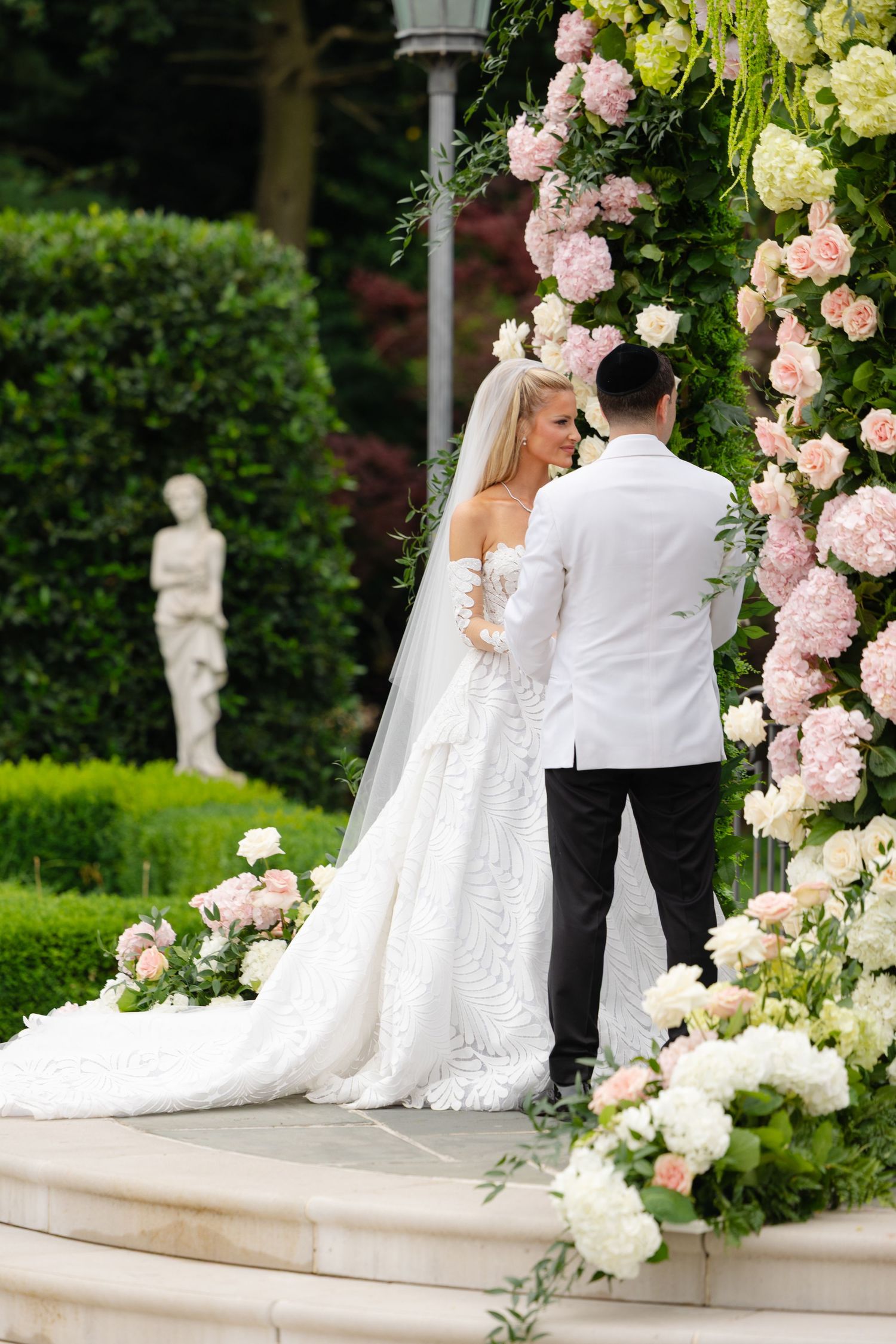 A bride and groom stand under an elegant floral arch with pink and white roses during their outdoor garden ceremony.