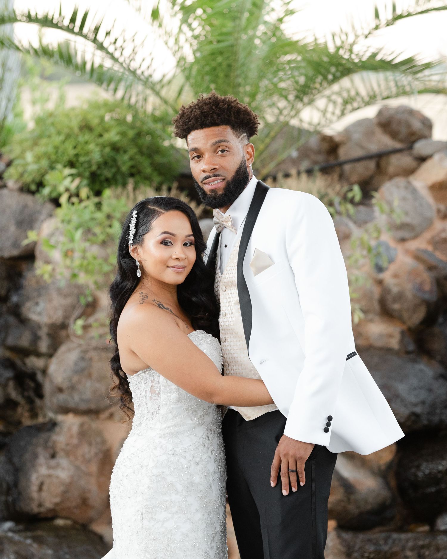 A couple poses together for a wedding photo in front of a stone wall and palm trees.