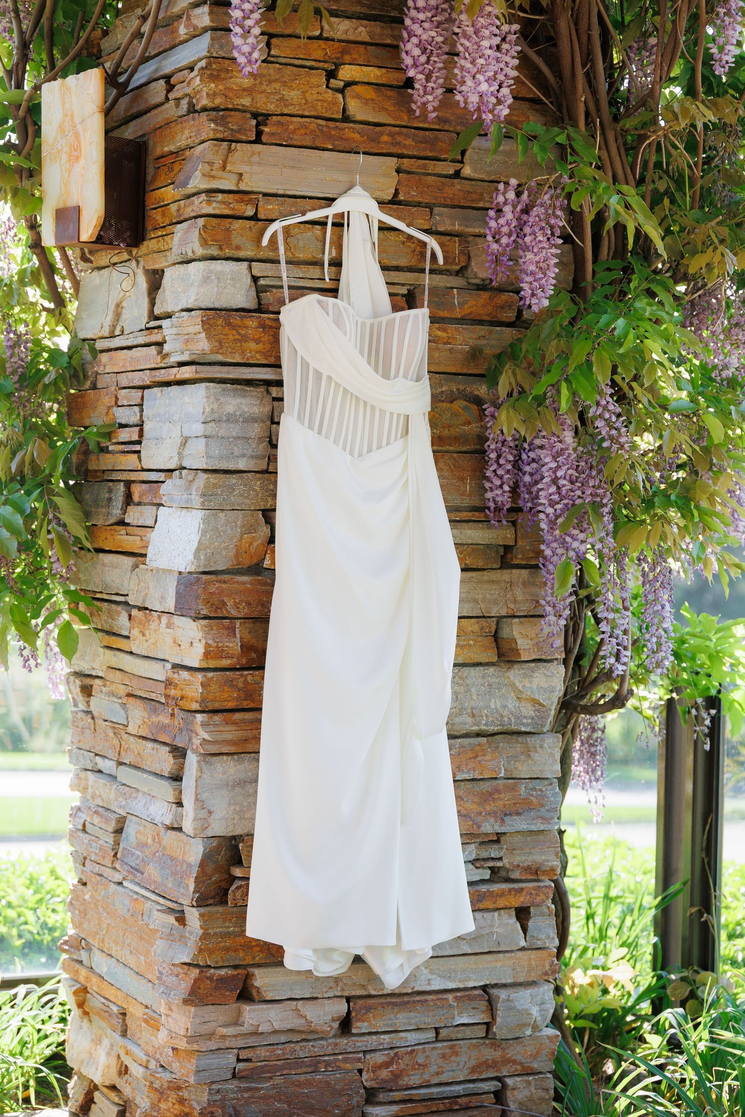 White vintage clothing hangs on wooden hangers against a rustic stone wall with blooming wisteria vines.