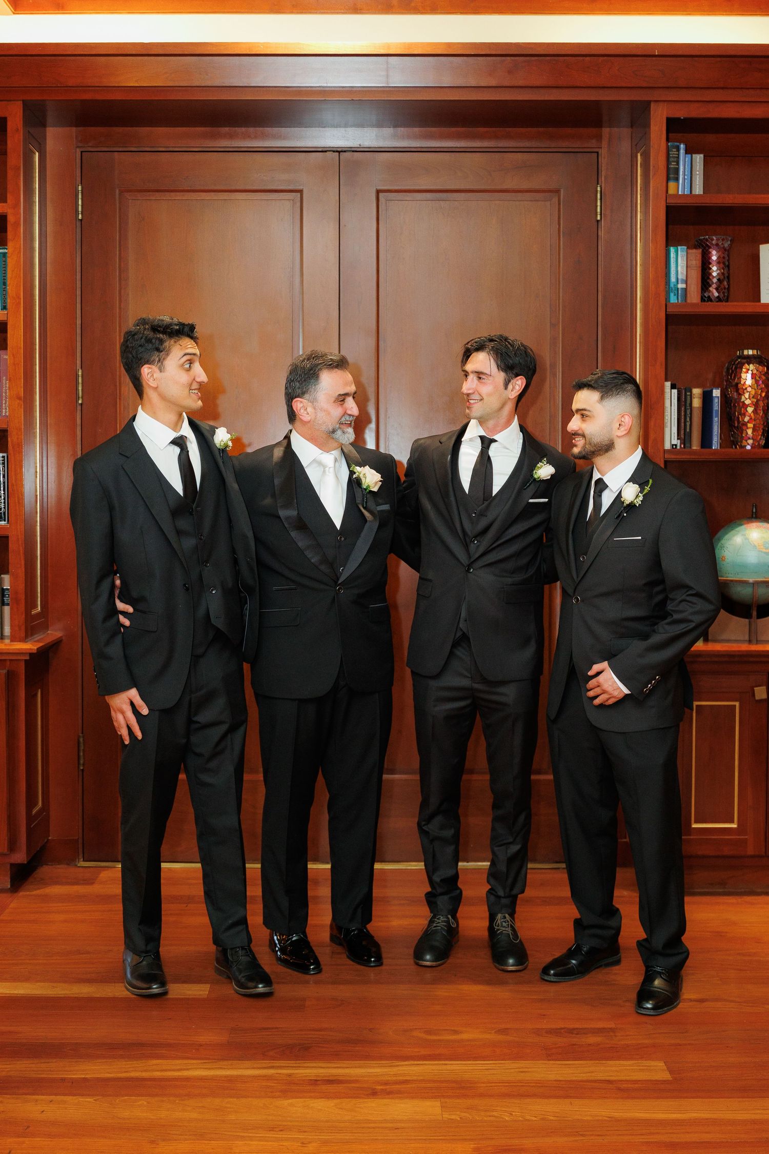 Groomsmen in black suits share a candid moment laughing together in a wood-paneled room.