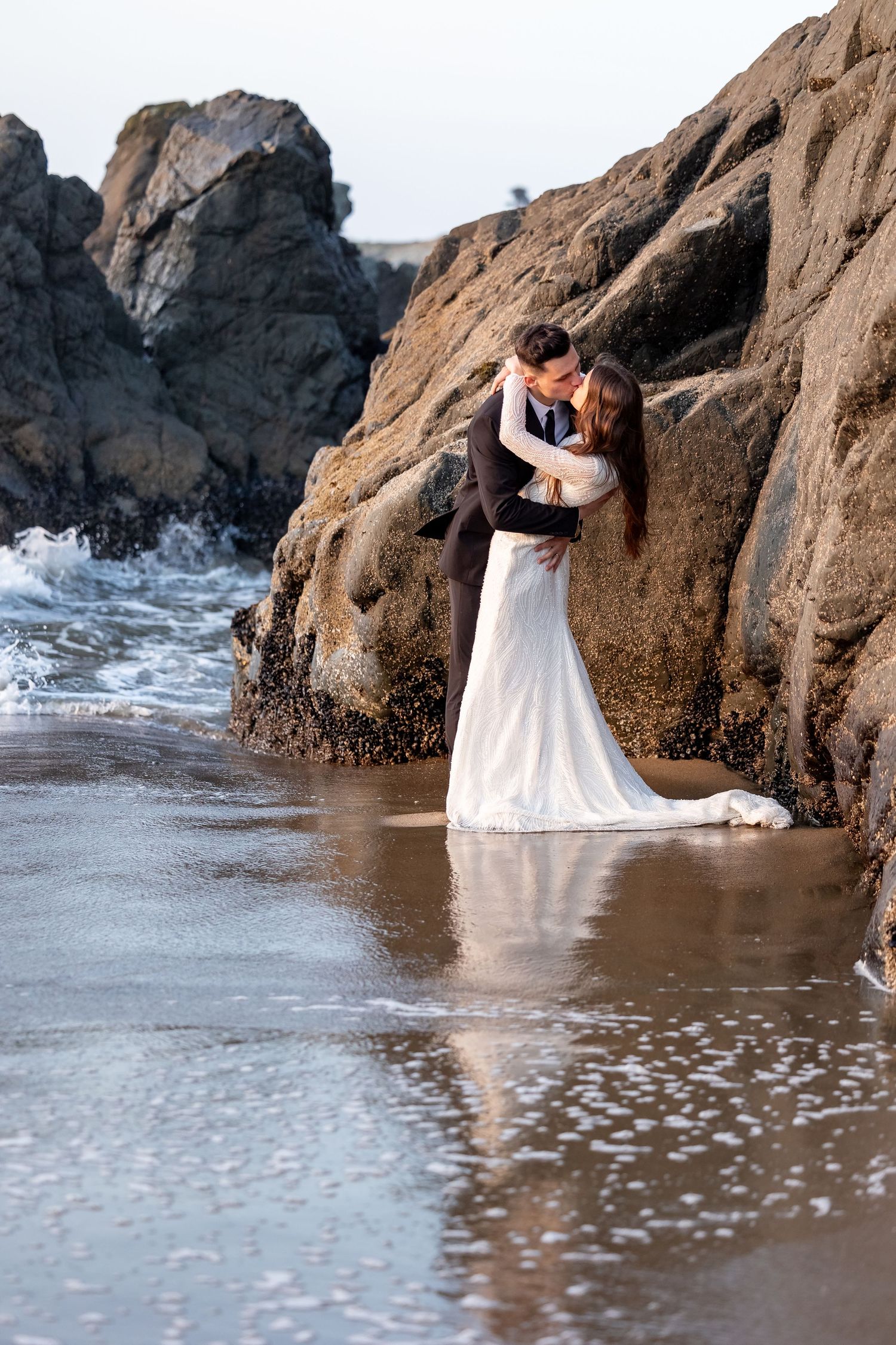 Romantic beach wedding photos with couple posing against dramatic coastal rocks.