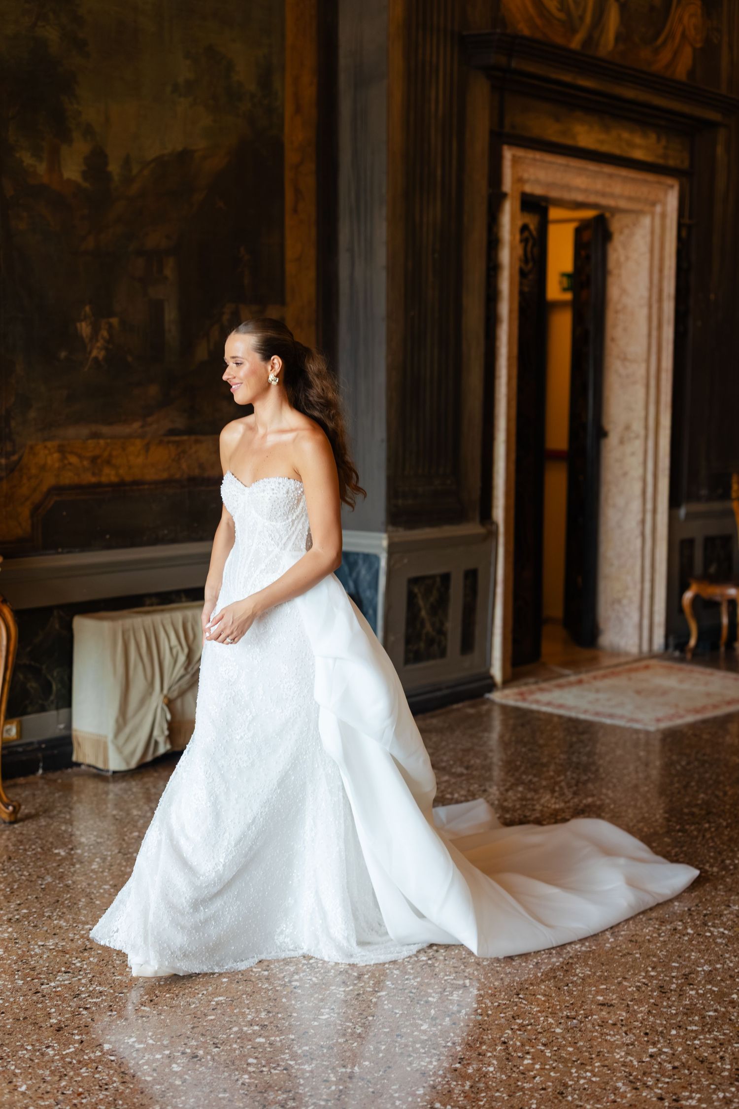 A bride in a flowing white wedding gown sits gracefully on a marble floor in an elegant historic interior.