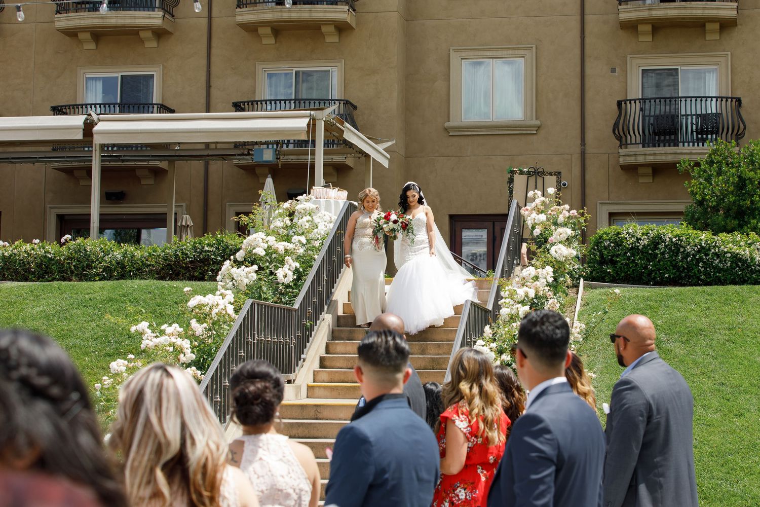 A wedding ceremony takes place on an outdoor staircase decorated with white floral arrangements at an elegant venue.