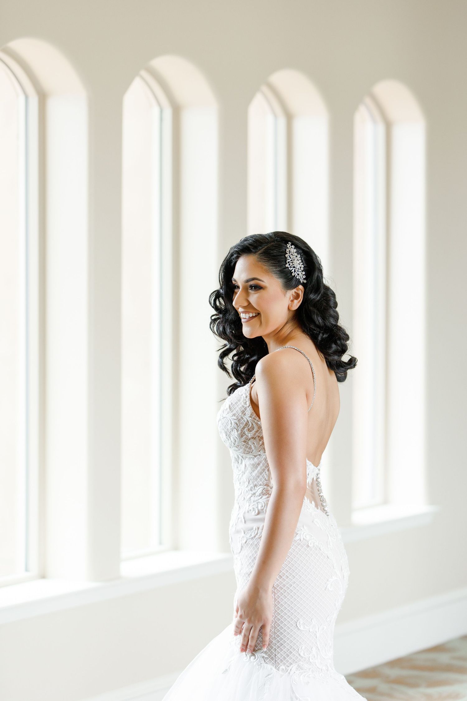 A bride in a white lace wedding dress poses elegantly in a bright sunlit hallway with arched windows.