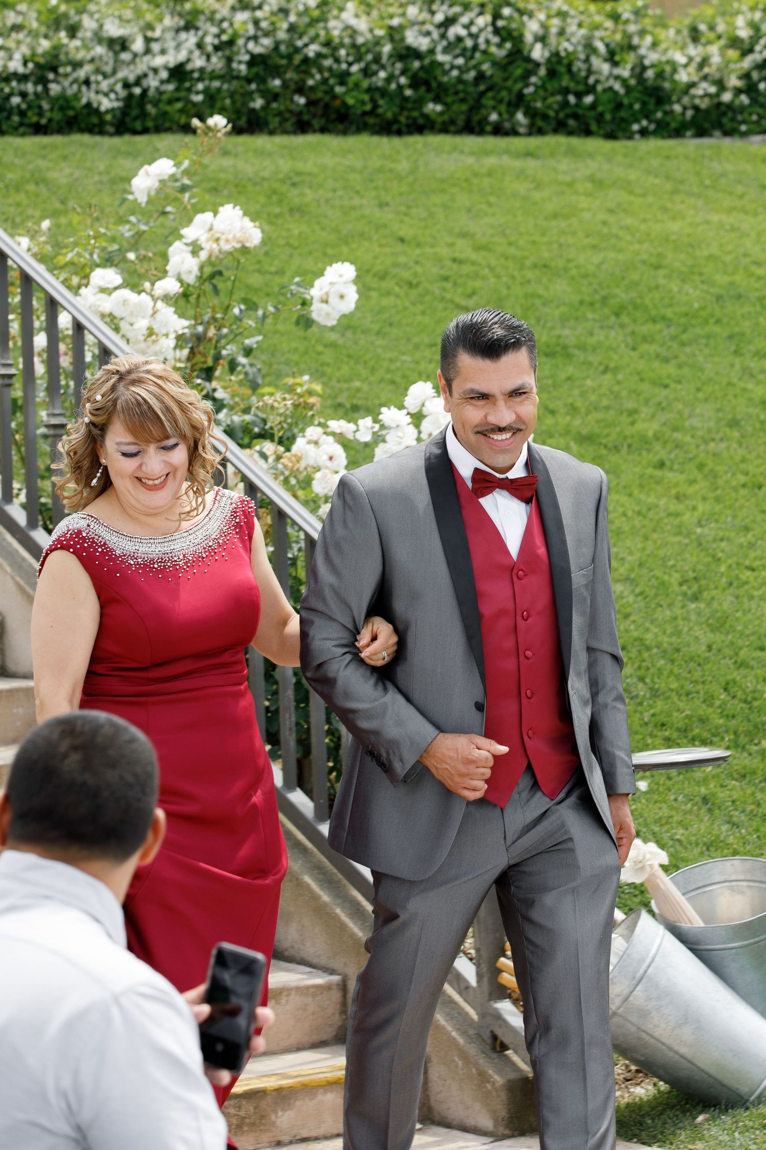 A couple in formal attire descends outdoor stone steps, with the woman in red and the man in a gray suit with red vest.