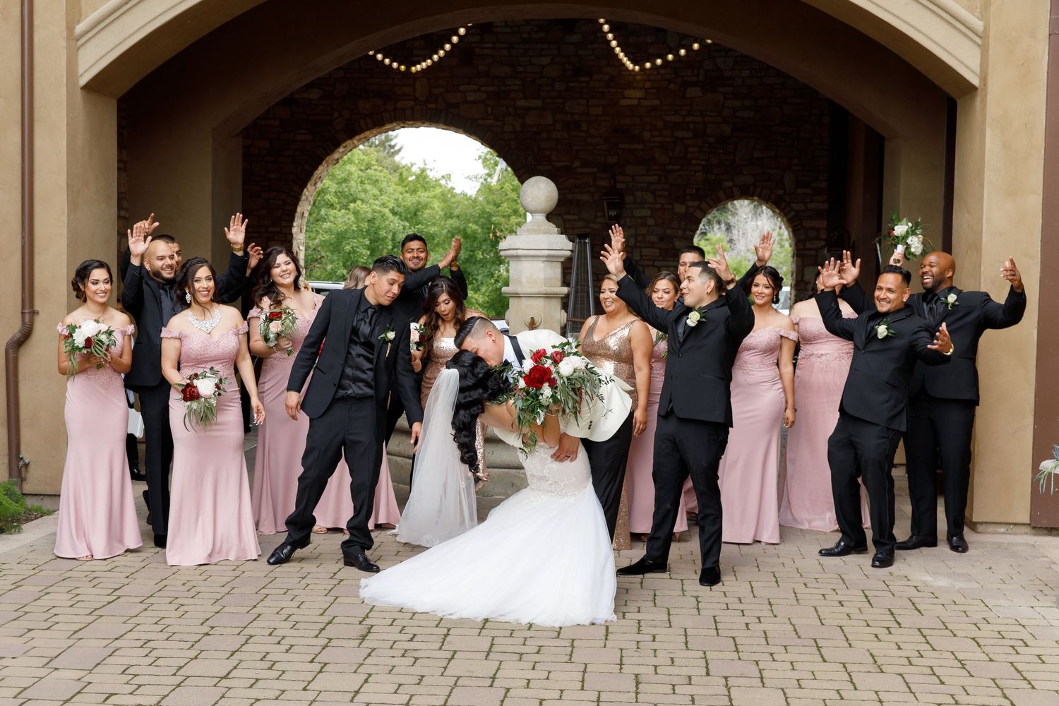 A wedding party poses joyfully under a stone archway, with bridesmaids in pink dresses and groomsmen in black suits.