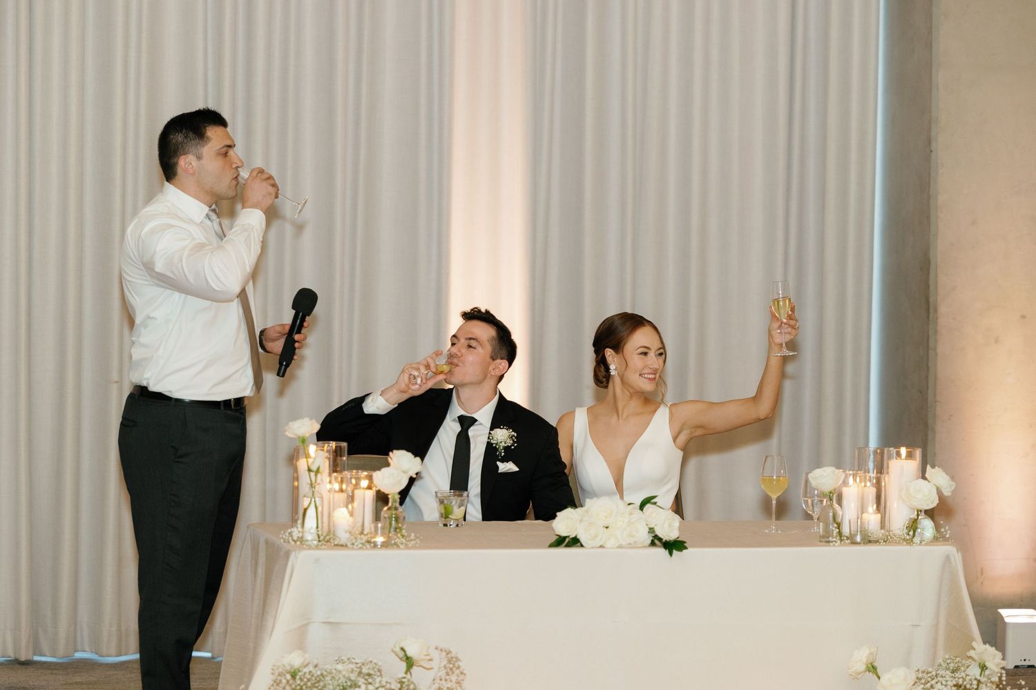 A group of people celebrating at an elegant wedding reception with white decor and candlelit tables.