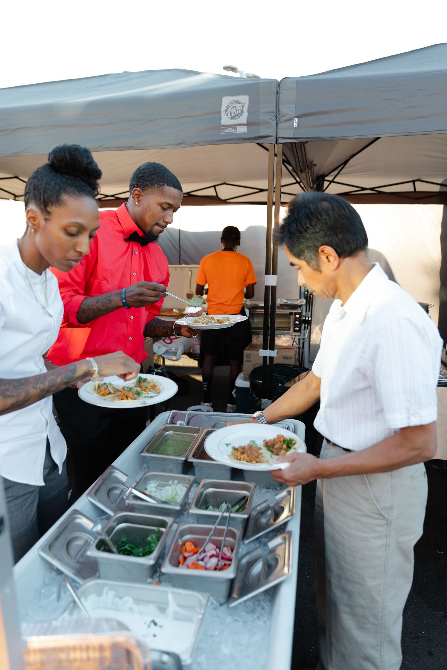Food service workers prepare and plate meals at an outdoor catering station with stainless steel serving trays.