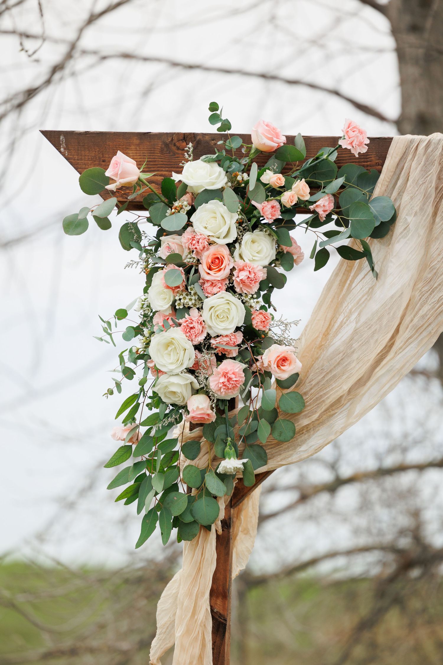 Beautiful floral arrangement with pink and white roses adorning a wooden arch with flowing champagne fabric draping.