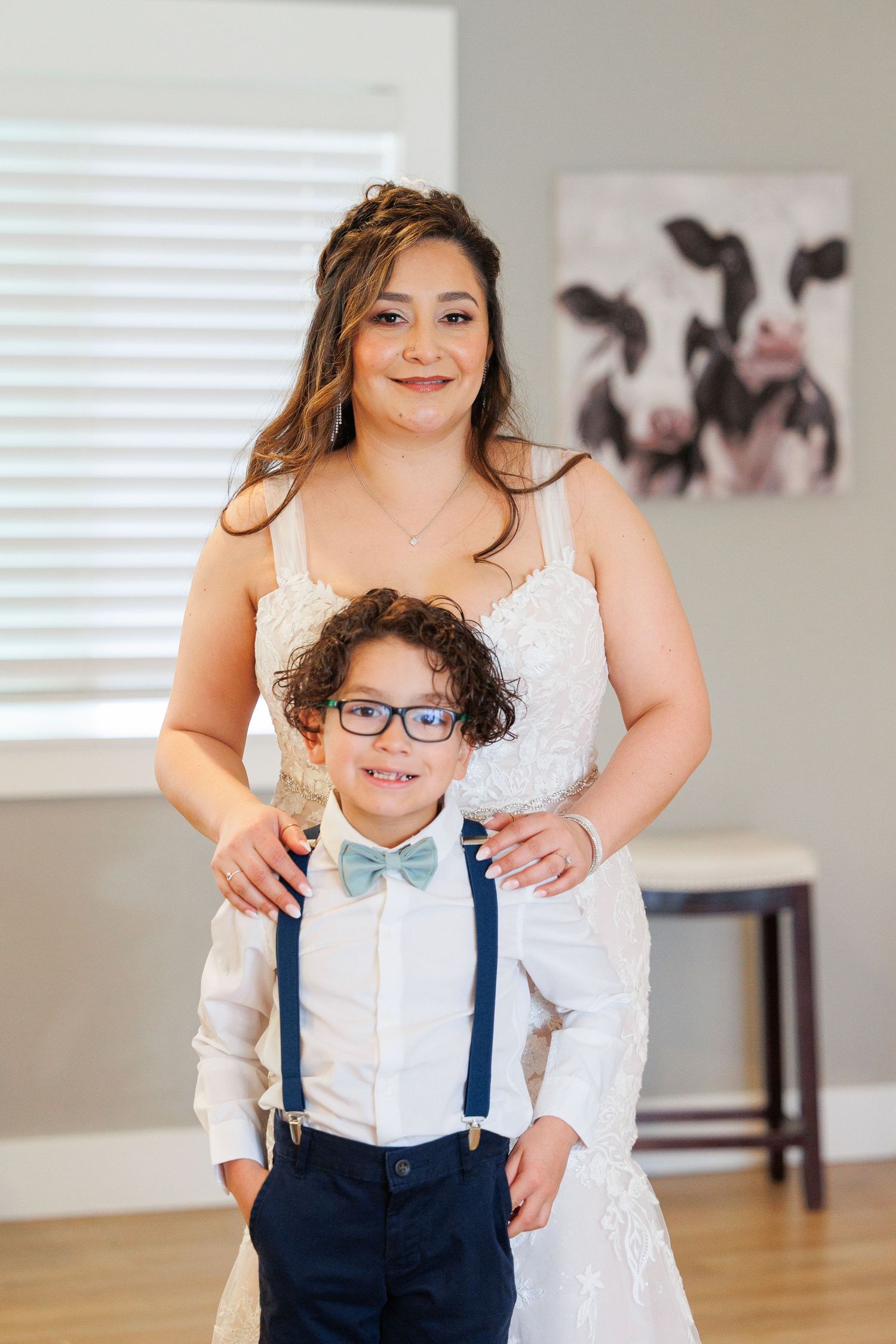 A young child in a white dress shirt, bow tie and navy suspenders poses with someone in a white dress indoors.