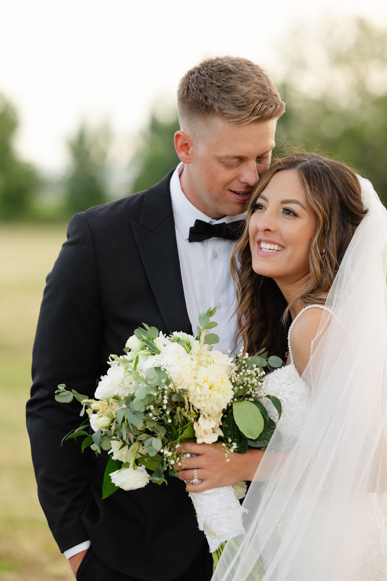 Romantic wedding couple embraces outdoors with white floral bouquet and classic black tuxedo.
