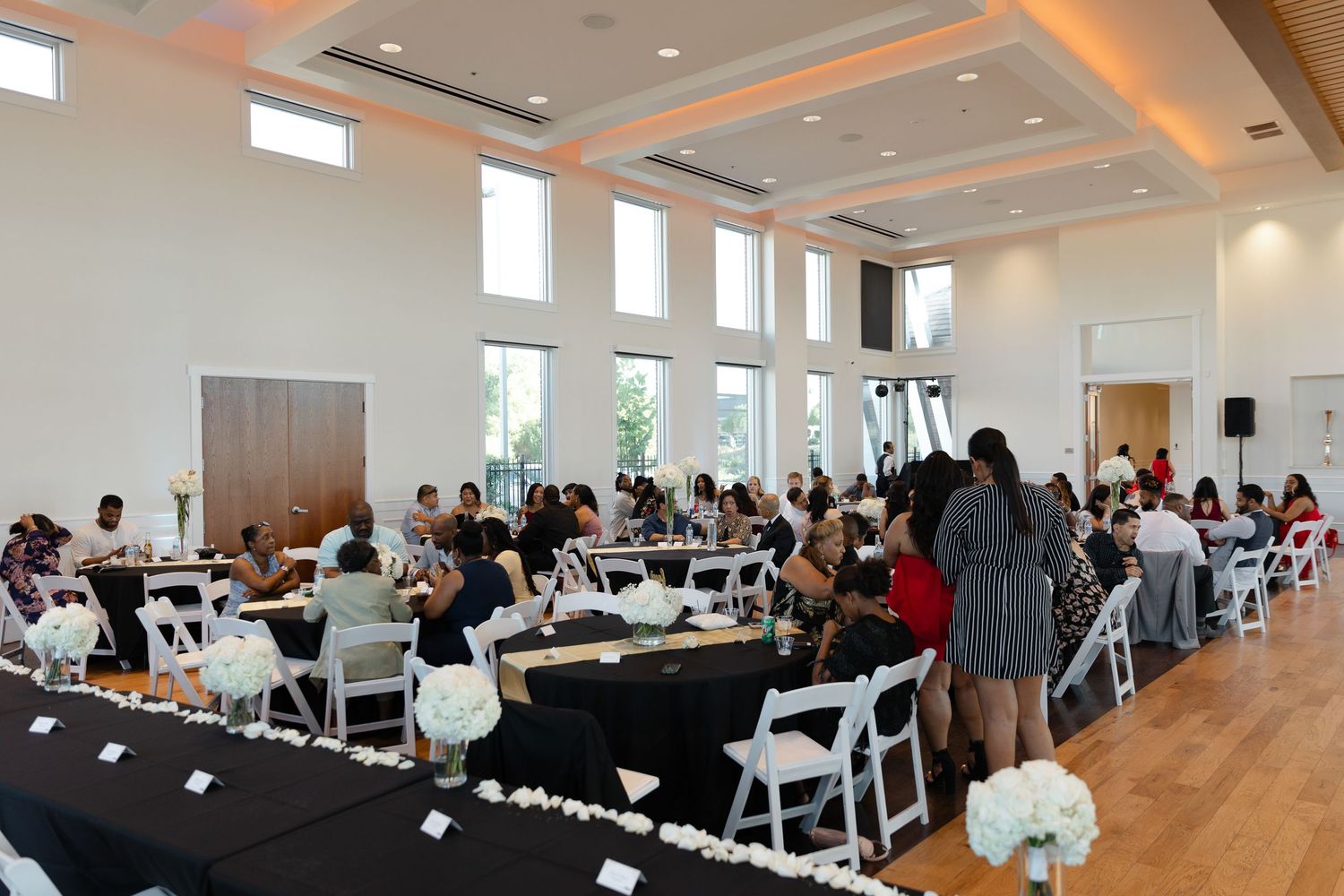 Large indoor wedding reception venue with white chairs, decorated tables and high ceilings with ambient lighting.