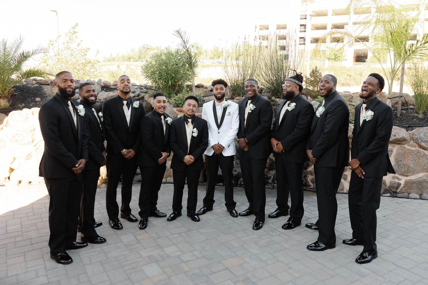 A group of groomsmen in black tuxedos pose in a curved formation outdoors at a formal wedding event.