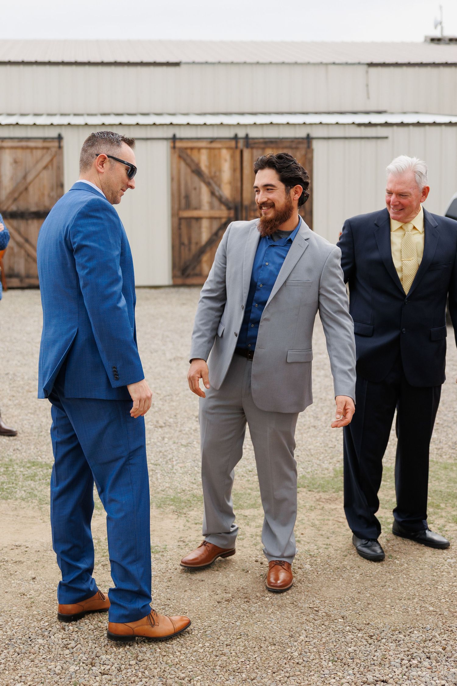 A group of groomsmen in blue and gray suits showing off their brown leather dress shoes on gravel ground.