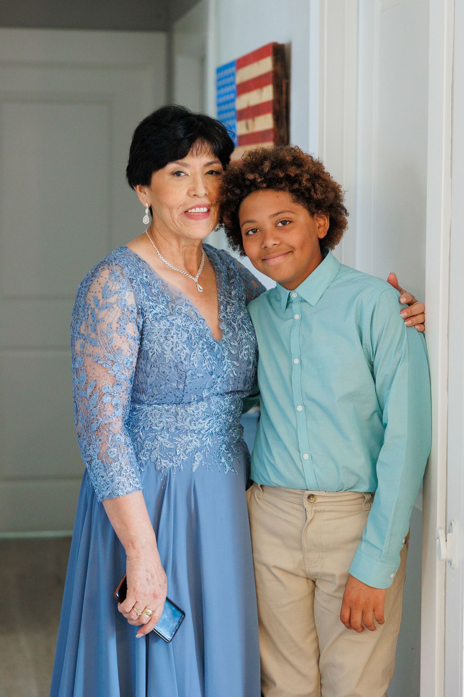 A woman in a blue lace dress poses with a young person wearing a mint green shirt and khaki pants against a white wall.