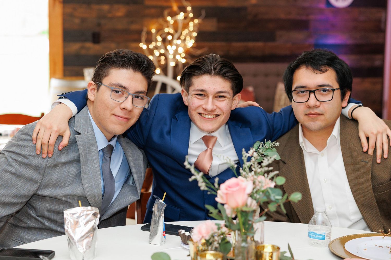 Three friends in suits smile together at a formal event with floral centerpiece and glowing lights in background.
