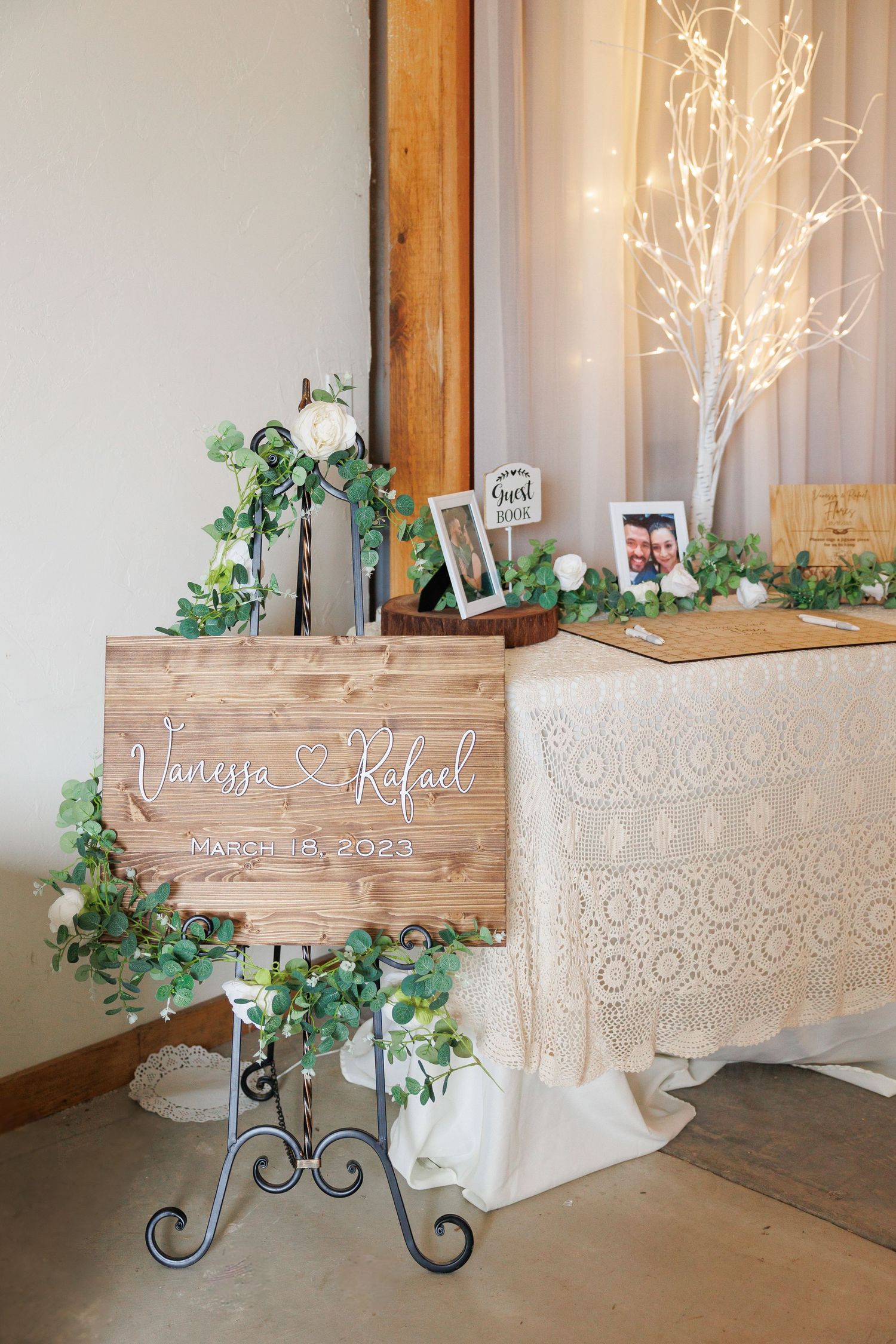 Elegant wedding cake display with lace-patterned tablecloth, greenery accents and illuminated branches in rustic venue.