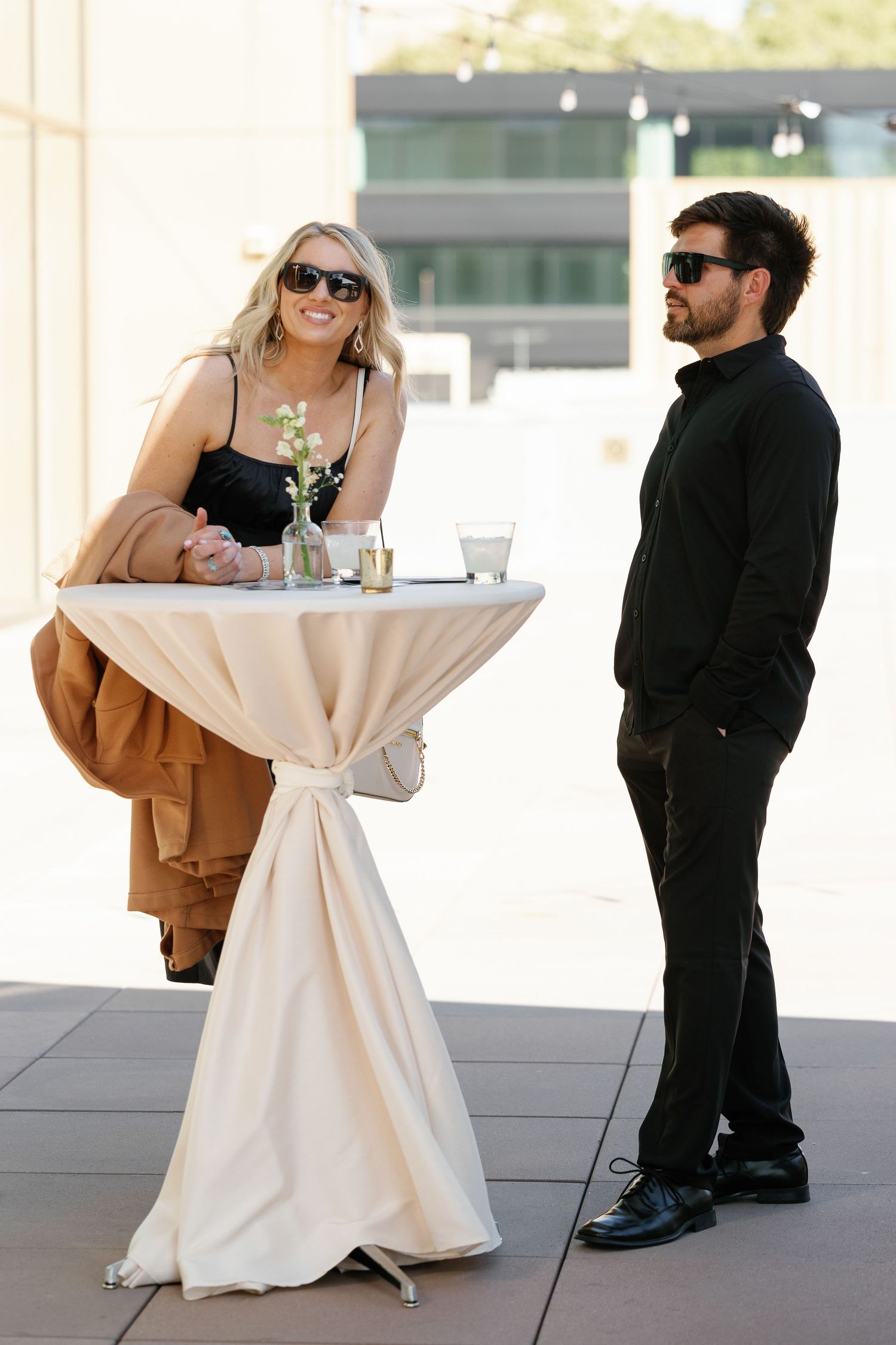 Two people in sunglasses chat while standing at a tall cocktail table with white tablecloth at an outdoor event.