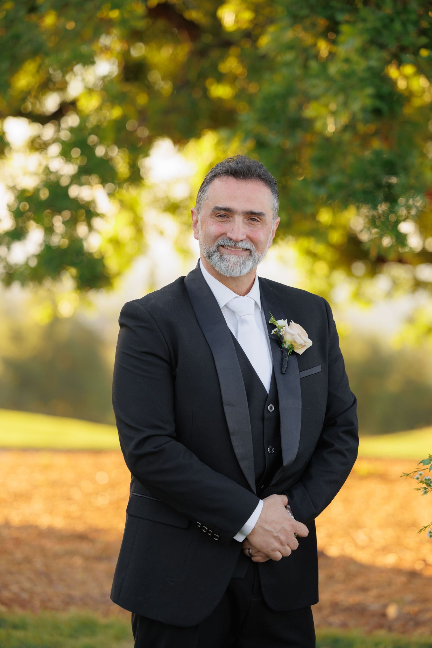 A groom in a black tuxedo and white tie stands outdoors in warm golden sunlight during a wedding photoshoot.