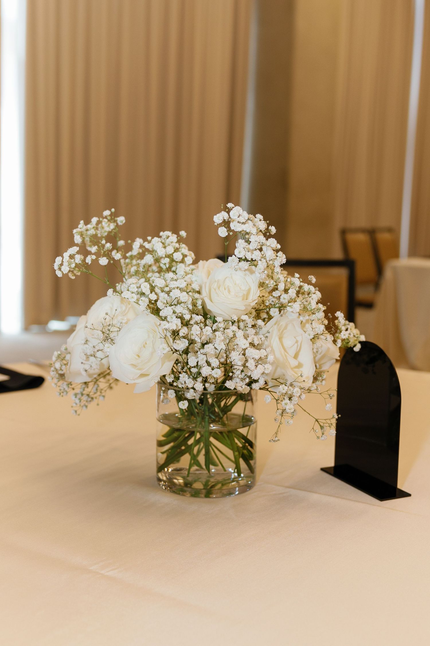 Elegant white roses and baby's breath centerpiece in glass vase with black table number holder on white tablecloth.