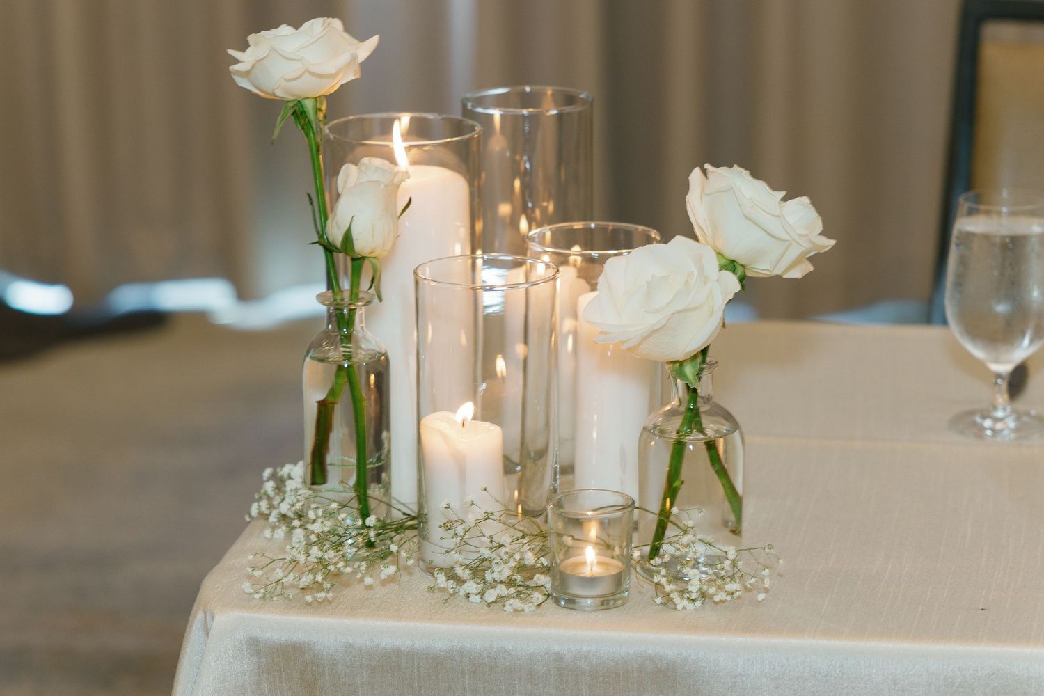Elegant wedding reception table setting with white roses, glowing candles, and delicate glassware on cream tablecloth.