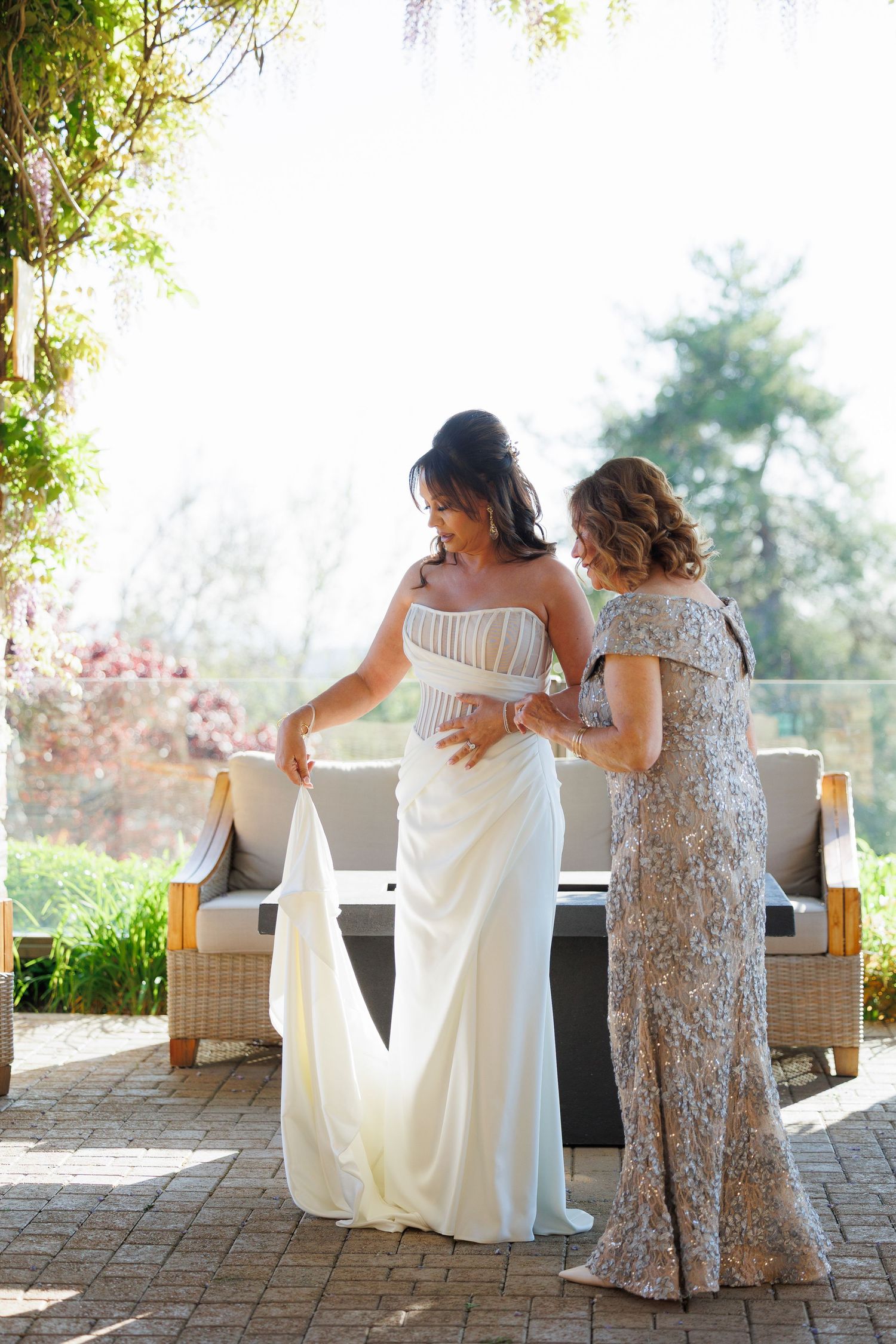 Two women in elegant dresses stand on a brick patio, one helping adjust the white wedding gown in natural outdoor setting.