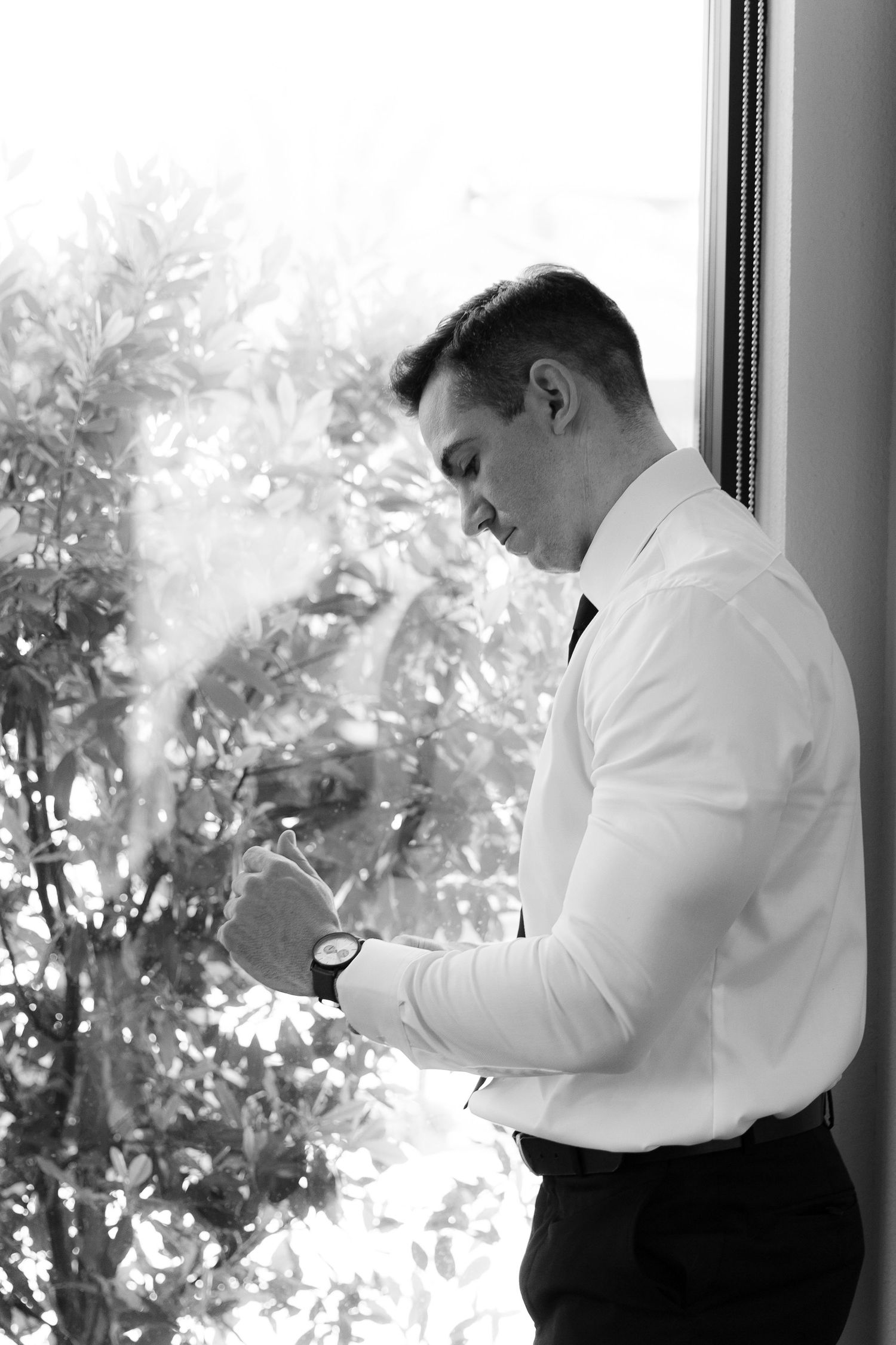 A person in formal attire stands by a window, looking down at something in their hand in a black and white photo.