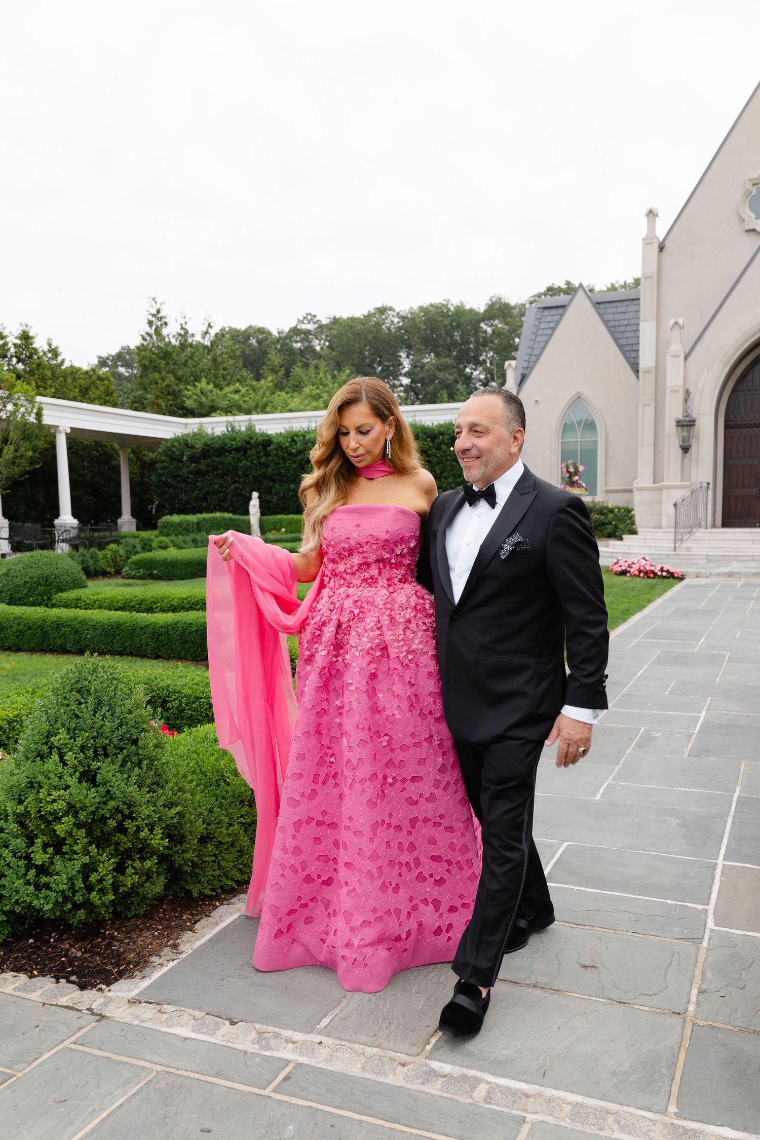 A couple poses outdoors in formal attire, with the woman wearing a flowing bright pink gown and the man in a black tuxedo.