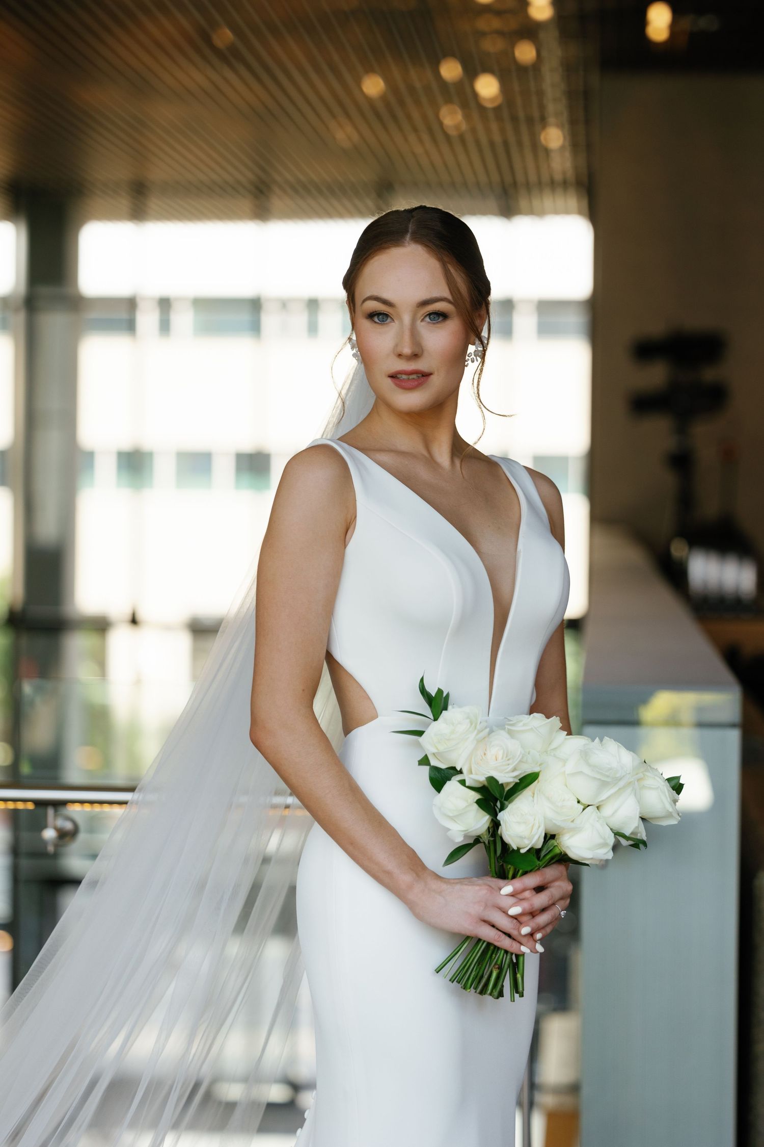Elegant bride in modern white wedding dress with plunging neckline and cutouts holds fresh white peony bouquet.