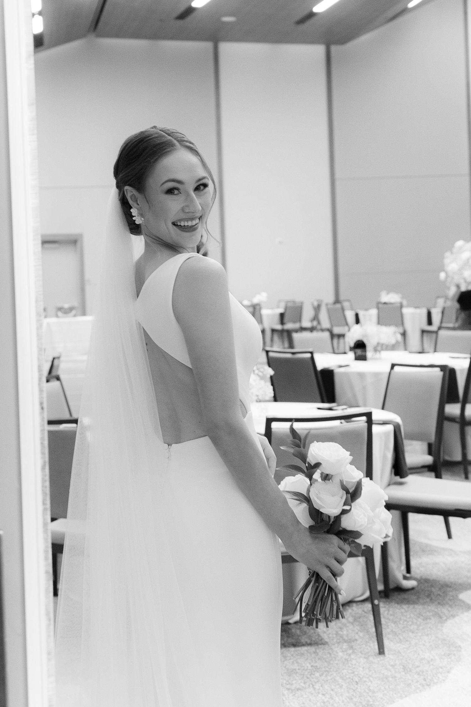 A bride in a white wedding gown smiles joyfully in an elegant reception venue with modern decor and lighting.
