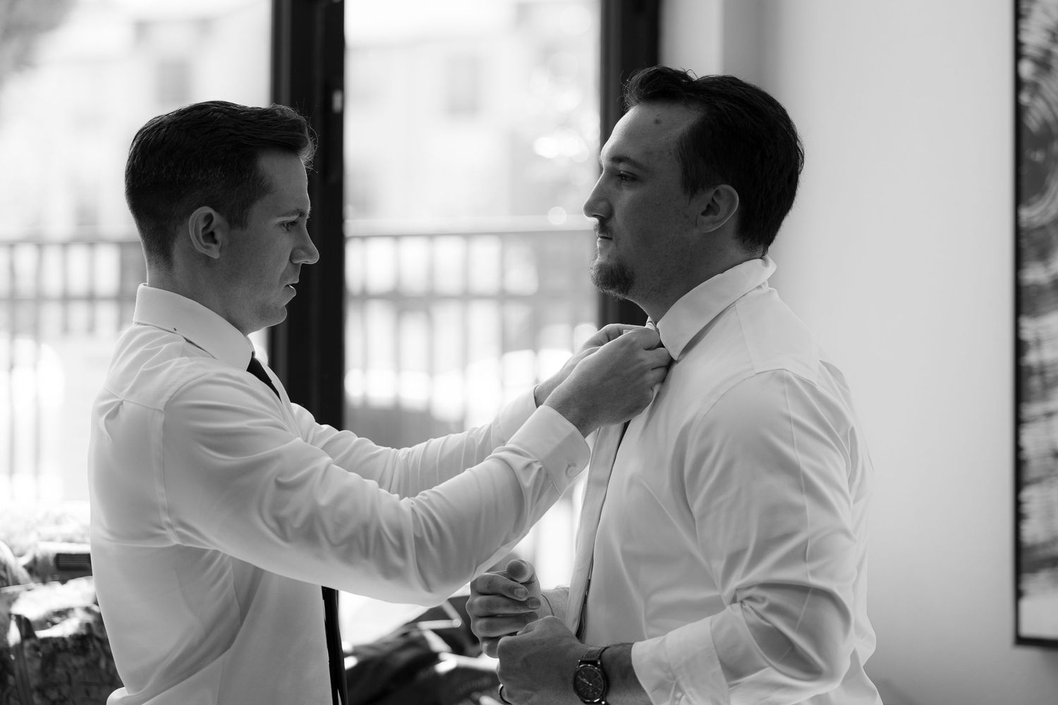 A black and white photograph shows one person helping another adjust their necktie in a formal getting-ready moment.