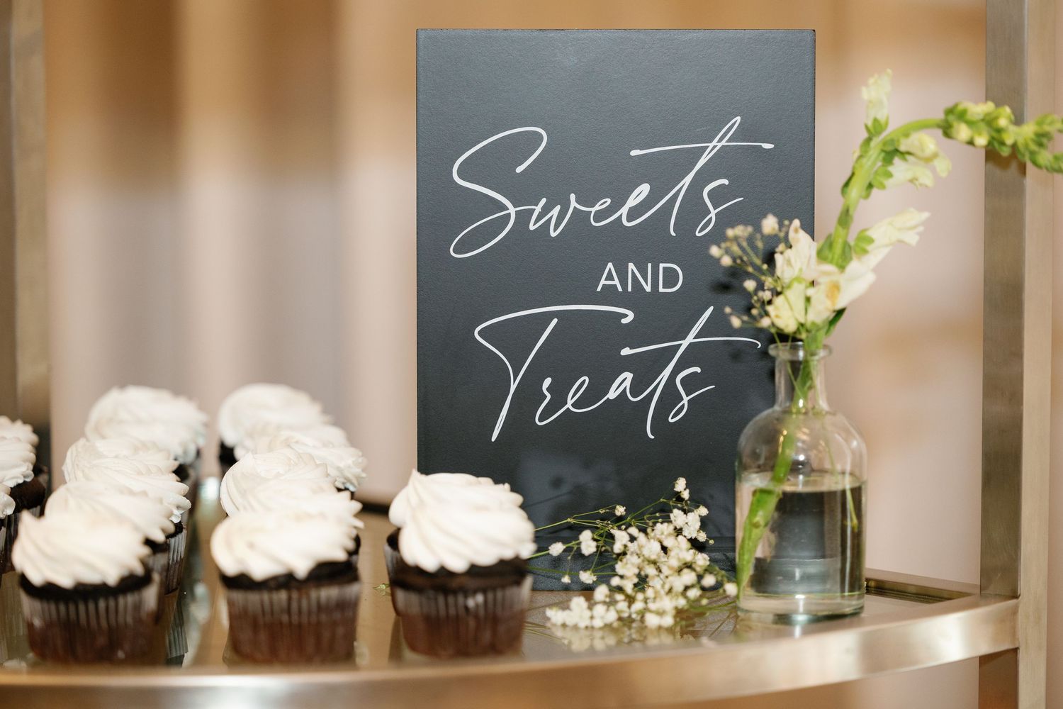A rustic wedding dessert display features chocolate cupcakes and chalkboard signs on wooden shelves with white flowers.