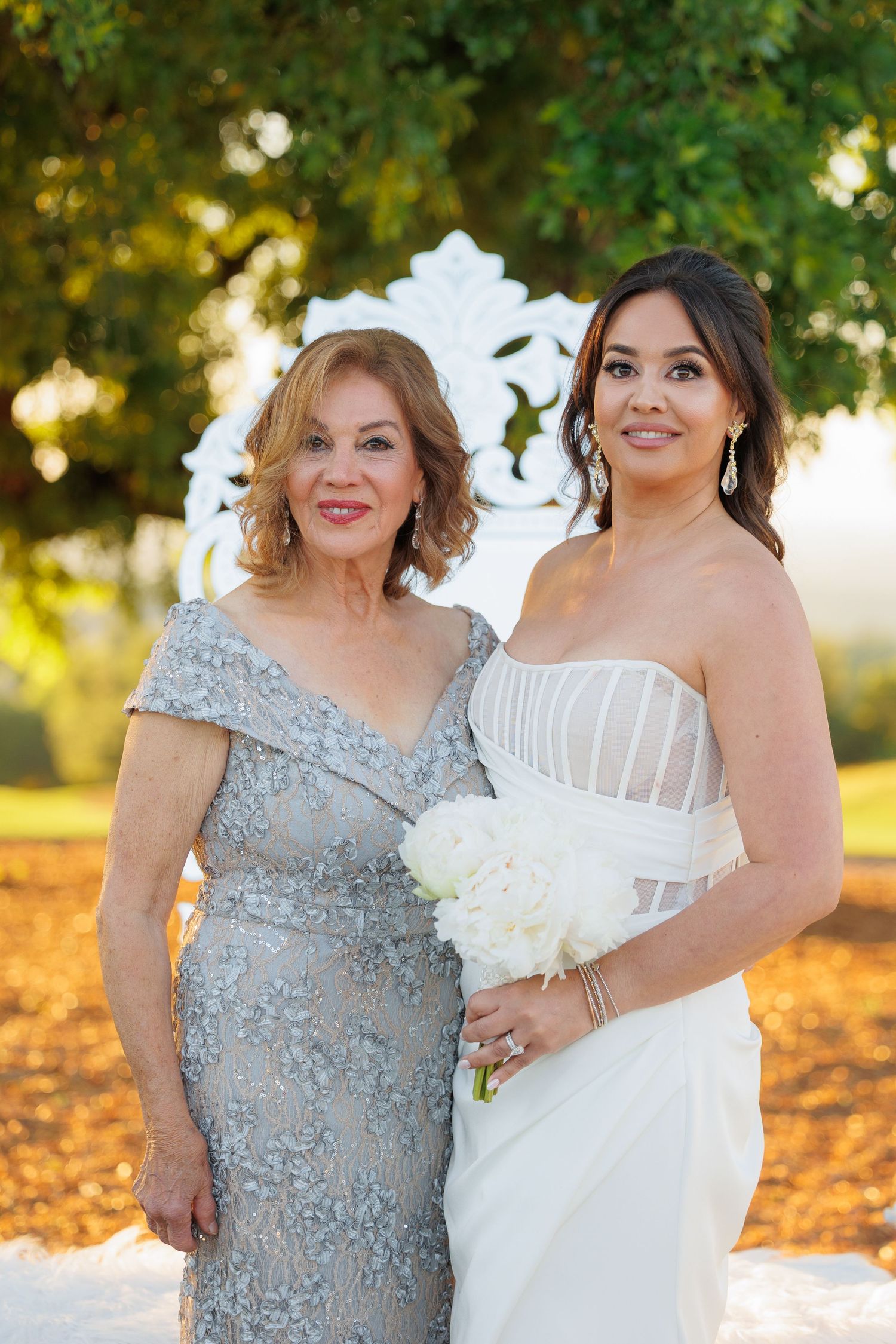 A pair of women in formal attire, one in a silver beaded gown and one in white, pose together outdoors during golden hour.