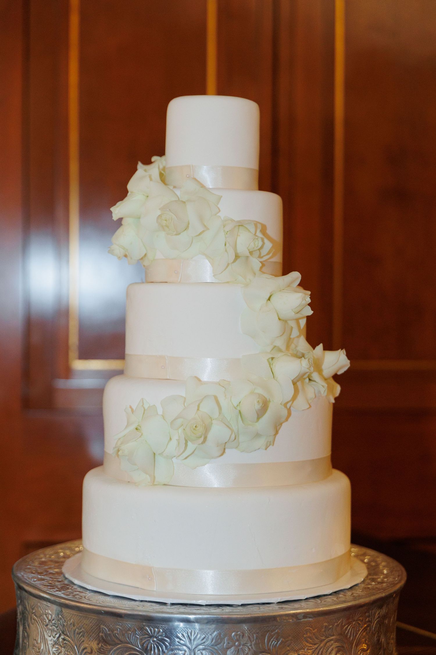 Elegant three-tiered white wedding cake with cascading white roses displayed on ornate silver cake stand.