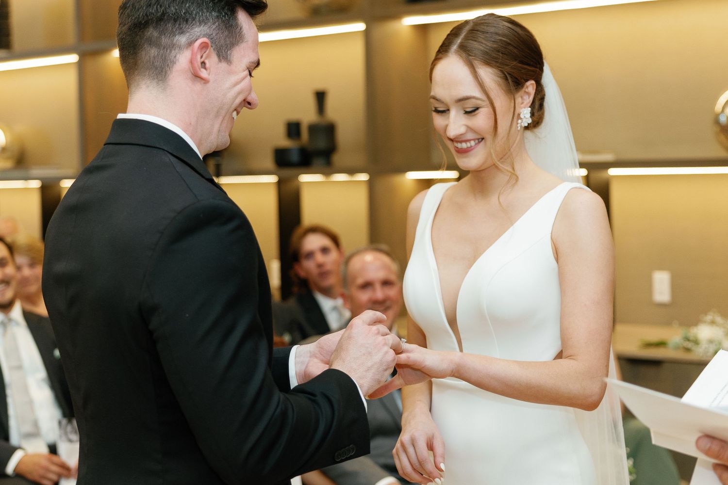 A couple exchanges rings during an intimate indoor wedding ceremony with guests looking on from their seats.