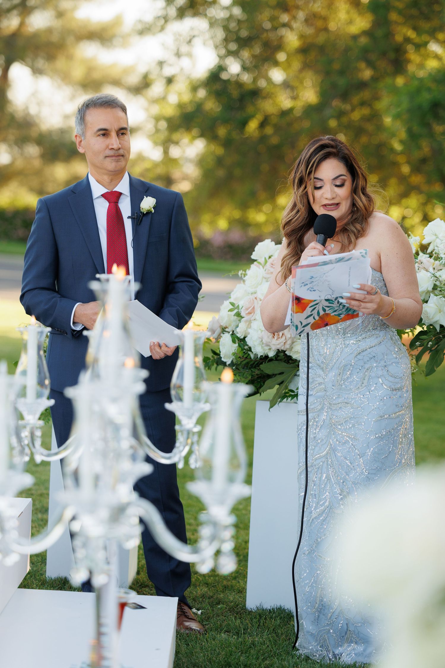 A couple in wedding attire interacts with a decorative white chandelier in an outdoor garden setting.