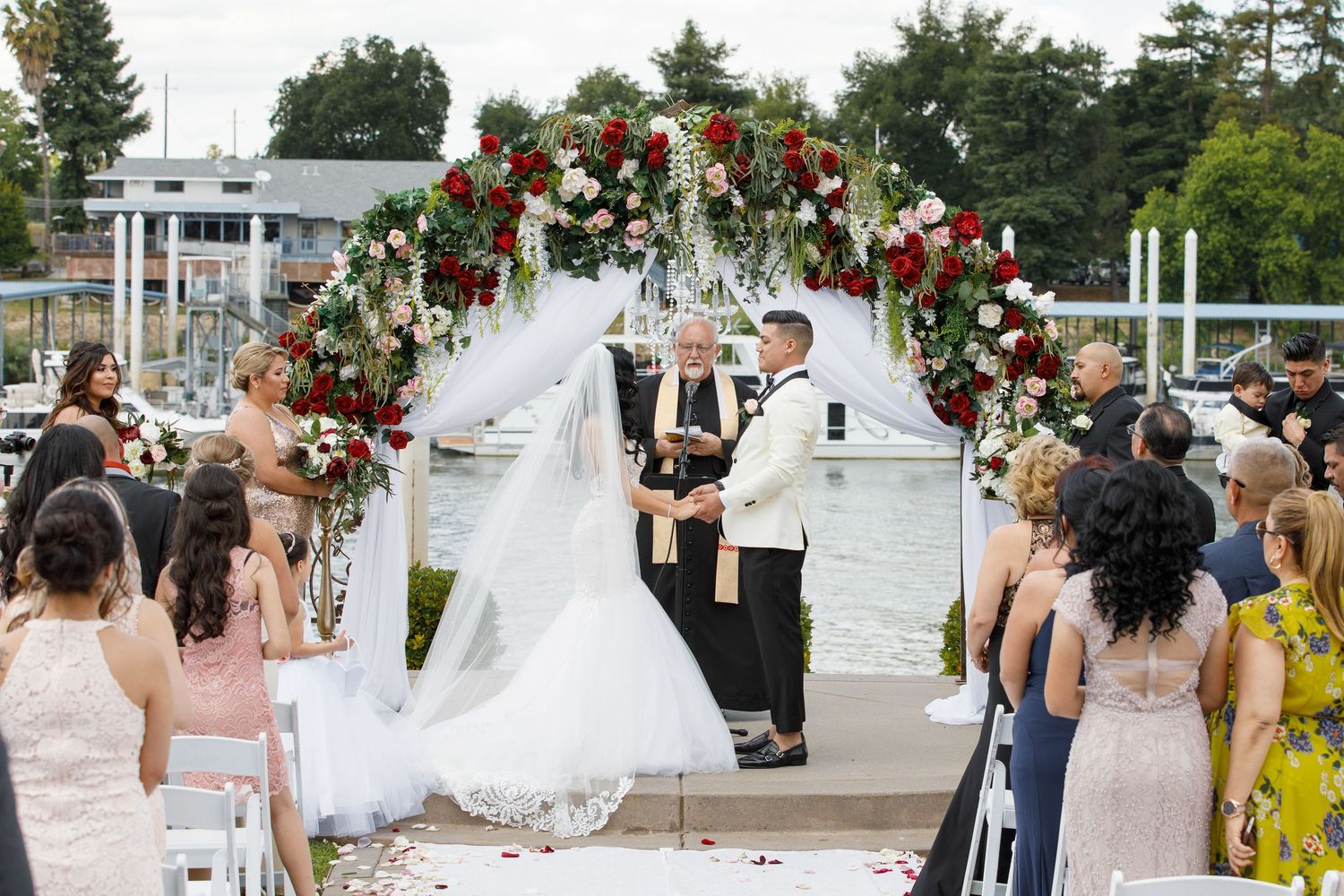 Wedding guests gather outdoors at an elegant venue decorated with red floral arrangements and white drapery.