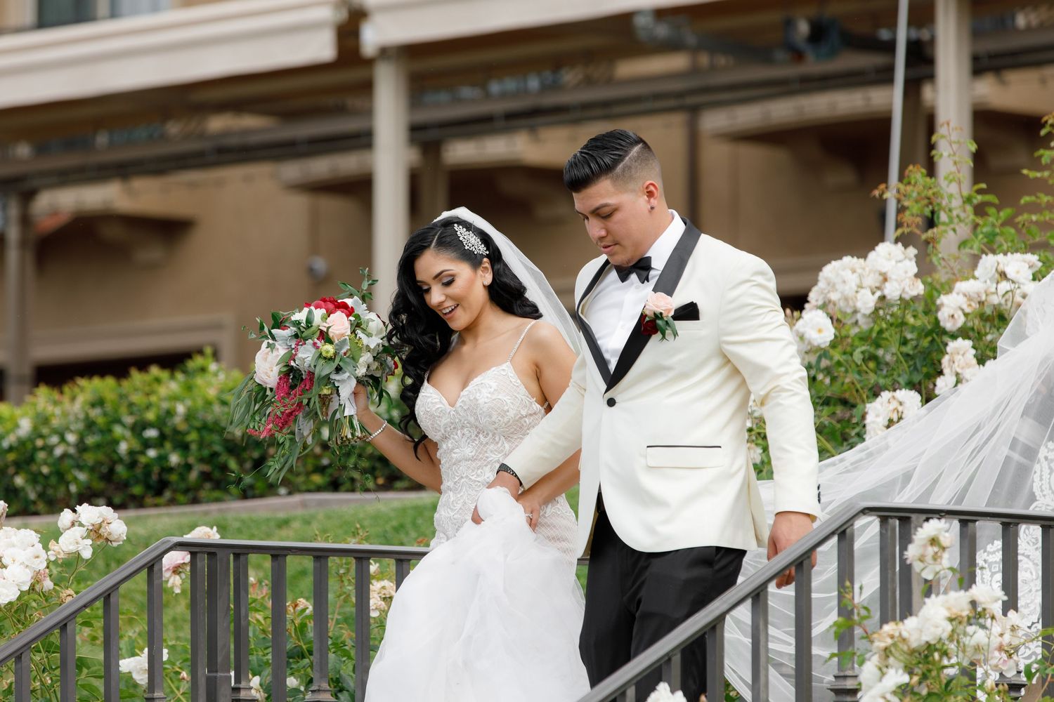 A bride and groom share romantic moments on outdoor stairs decorated with white floral arrangements.