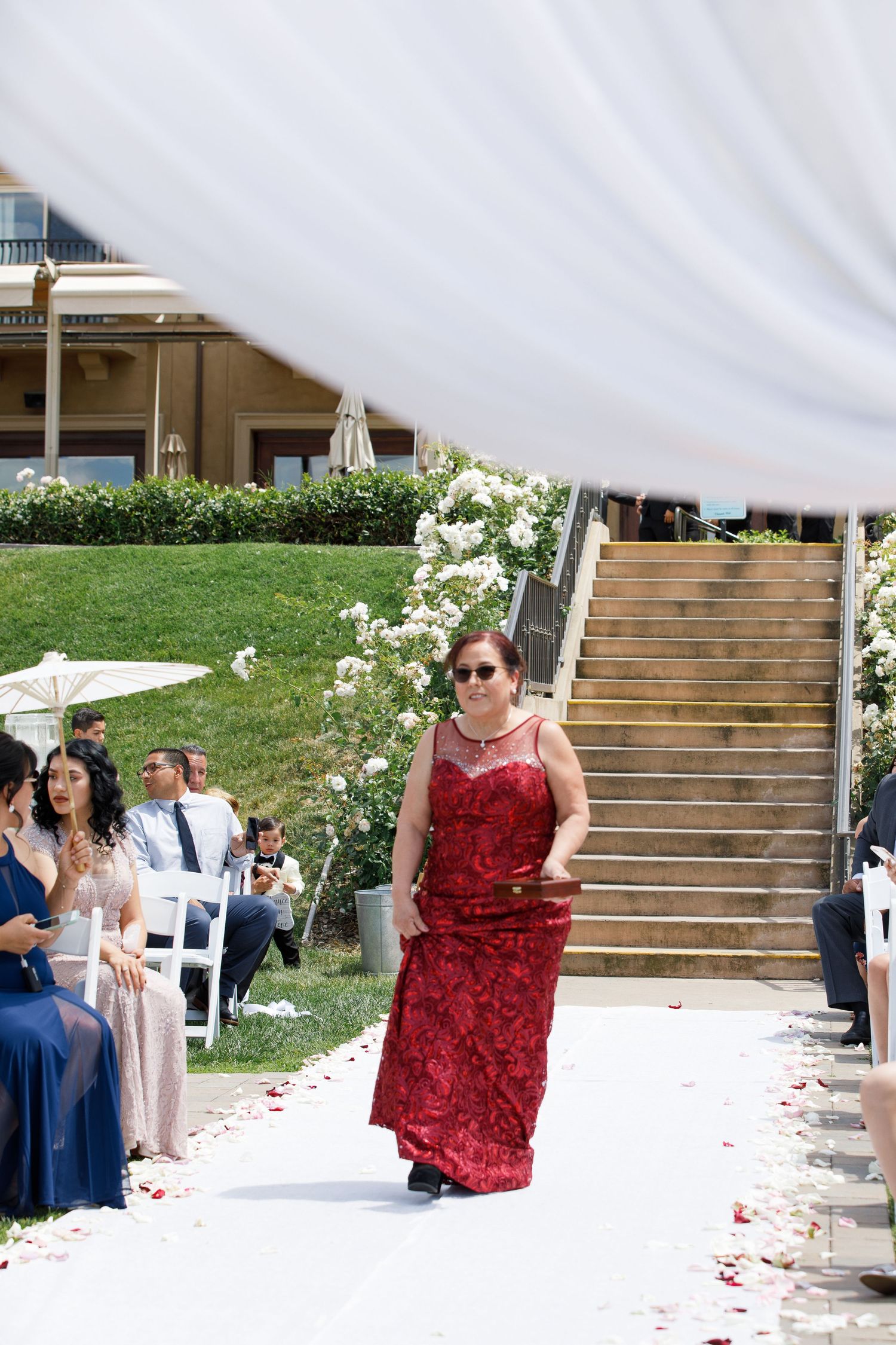 A wedding ceremony processional on a white aisle runner with red rose petals scattered, featuring an attendant in a red gown.